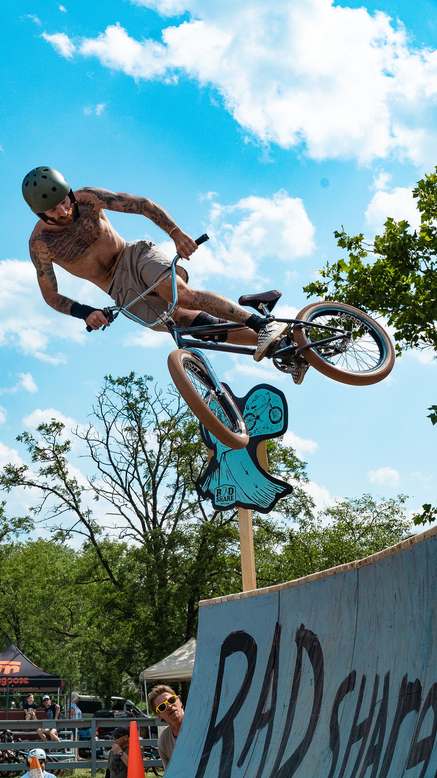 BMX rider executes trick over ramp at rad shack event with trees in background.