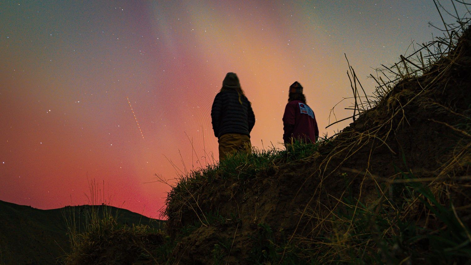 Silhouettes of two figures watching a vibrant sunset from a hilltop against a colorful sky.