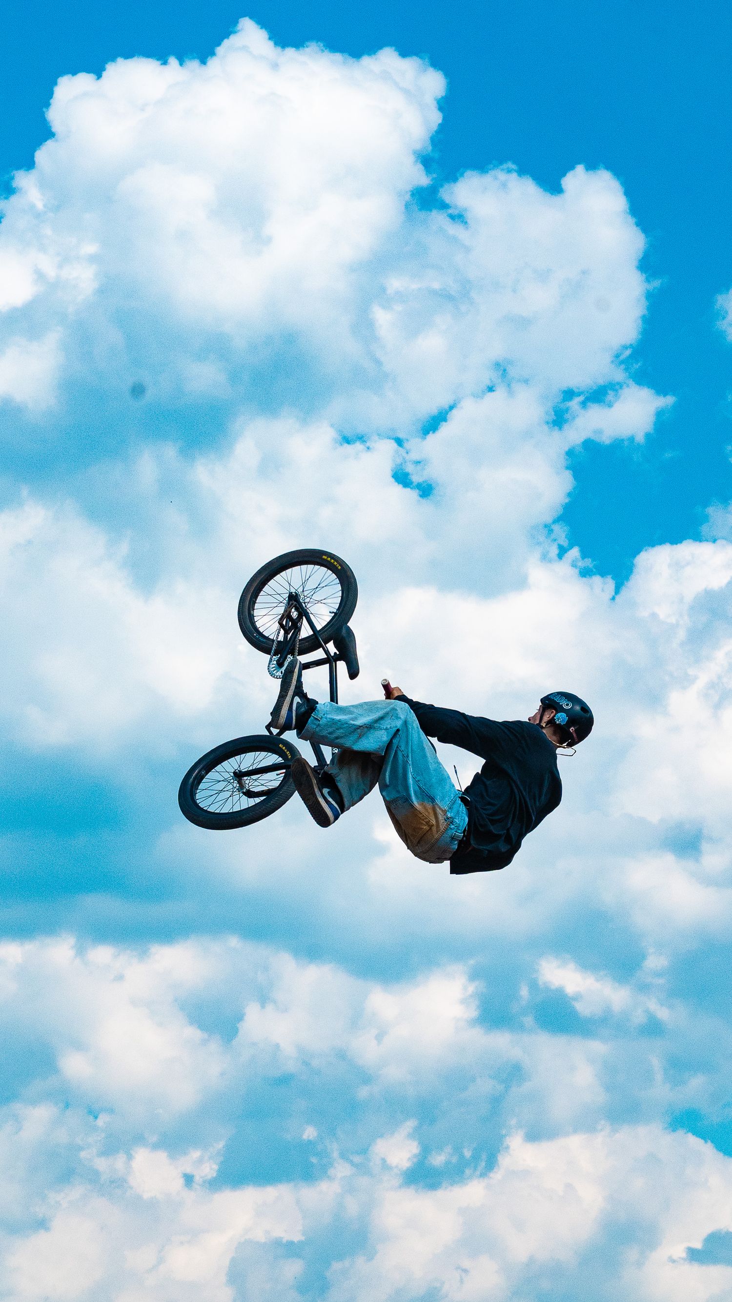 BMX rider performs a horizontal backflip trick against a bright blue sky with white puffy clouds.
