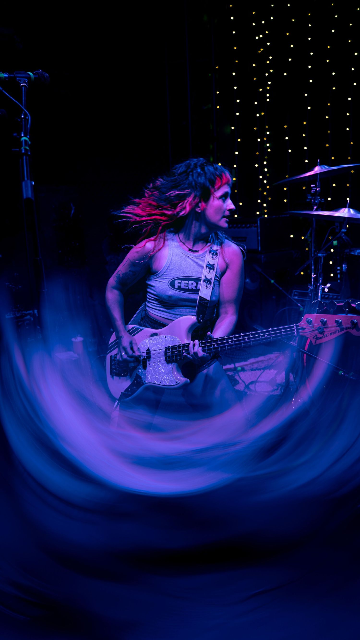 Bass player performs on stage with colorful blue and purple lighting effects creating dynamic swirling motion.