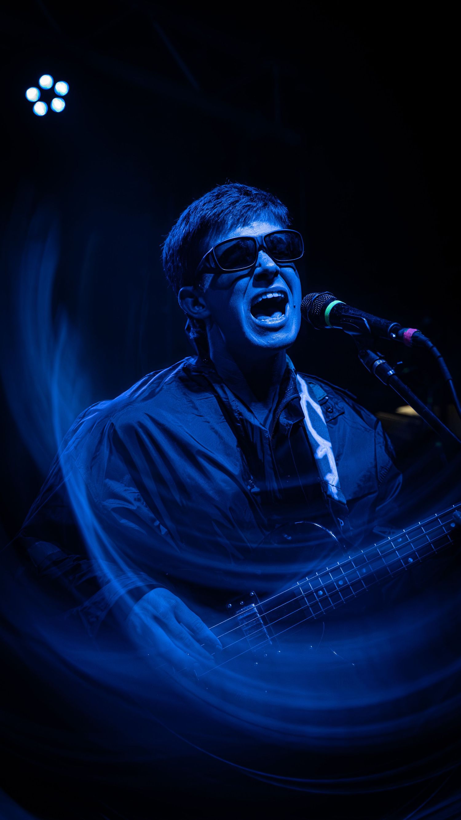 A musician performs on bass guitar against a dramatic dark blue stage lighting background.
