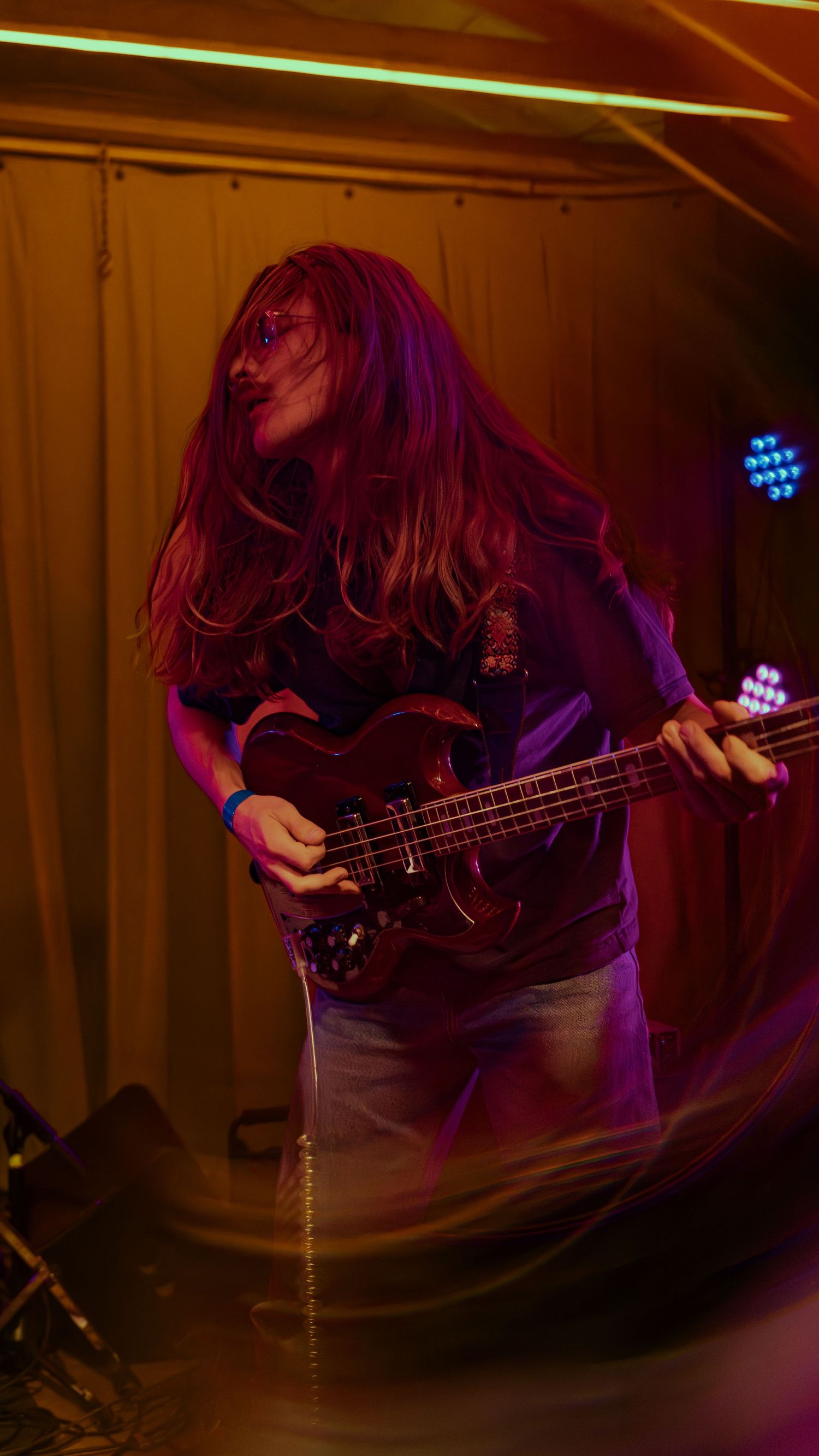 Silhouetted musician plays bass guitar on dimly lit stage with purple and blue lighting effects in background.