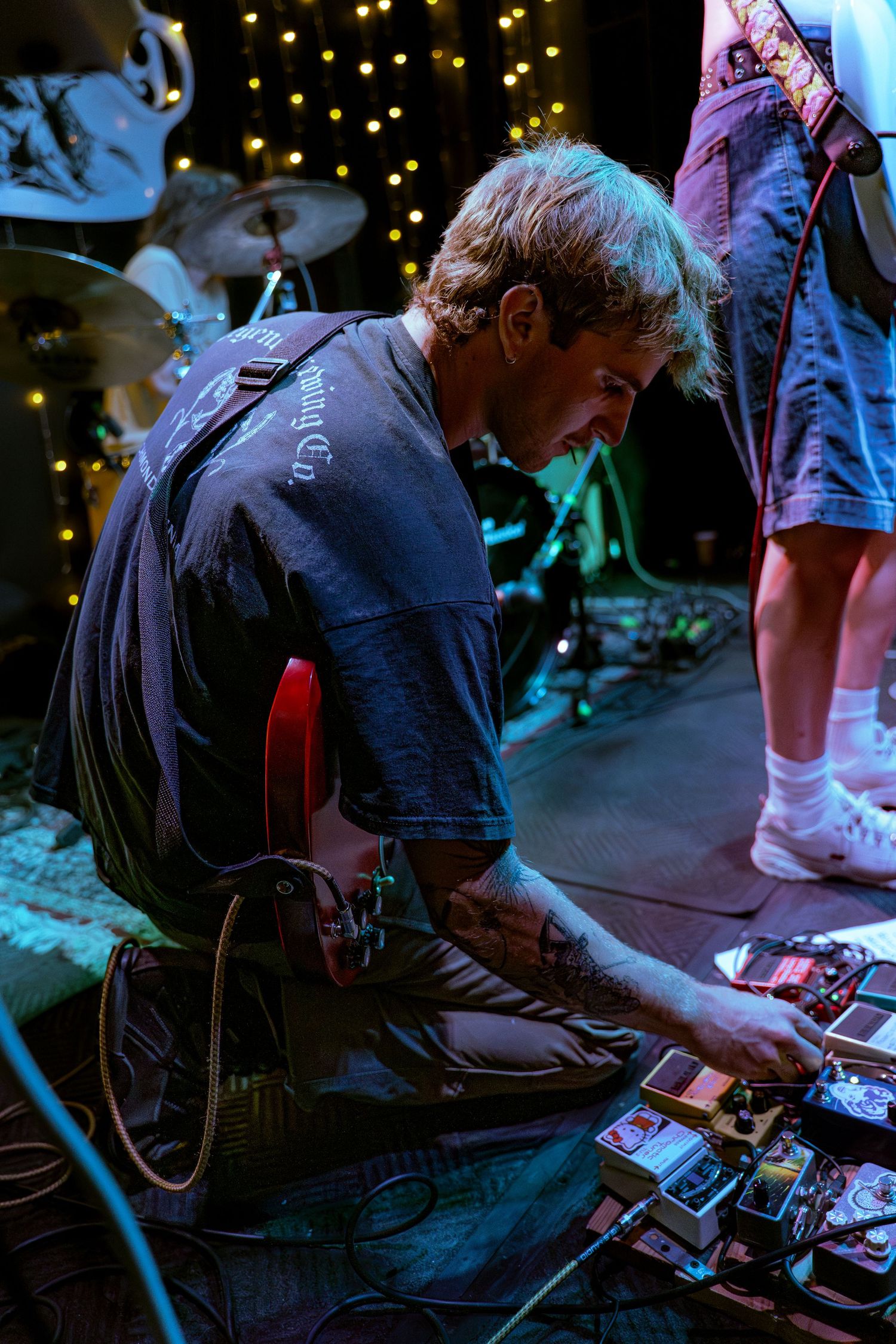 Musician bent over their pedalboard during a live performance with stage lights and bokeh in the background.