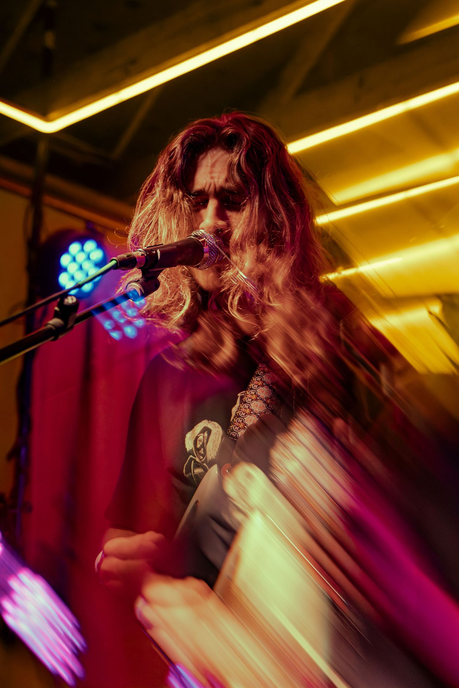 Concert performer with long hair stands on stage under dramatic yellow and purple lighting during a live music performance.