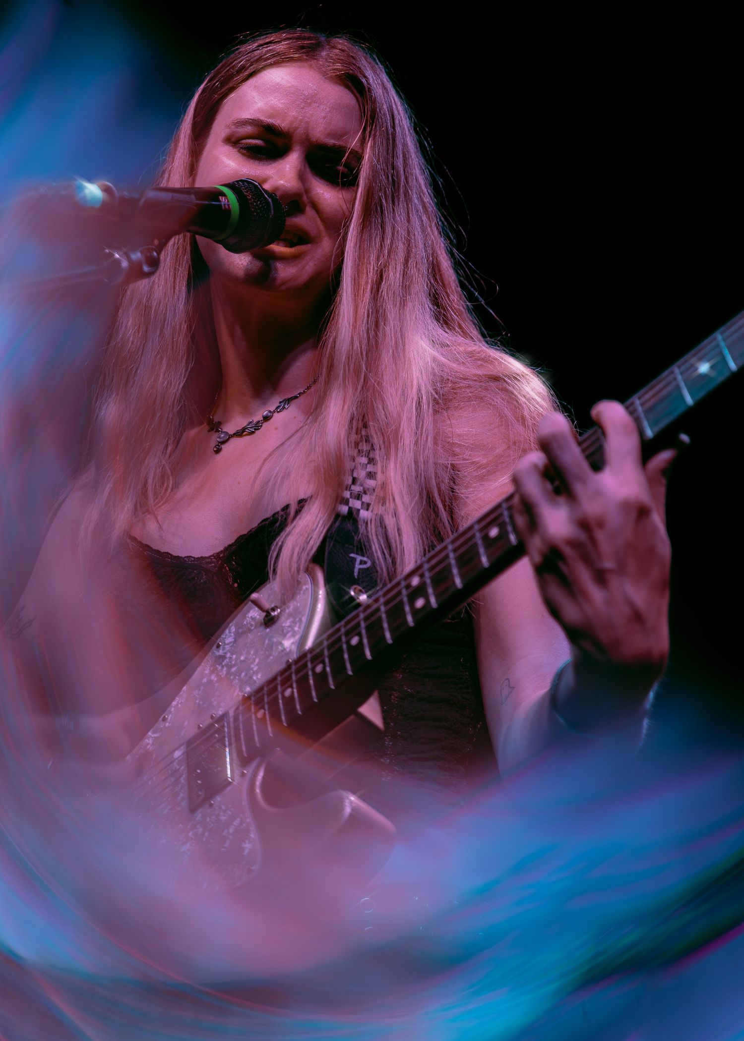Performer plays electric guitar and sings into microphone during concert with blue lighting effects.