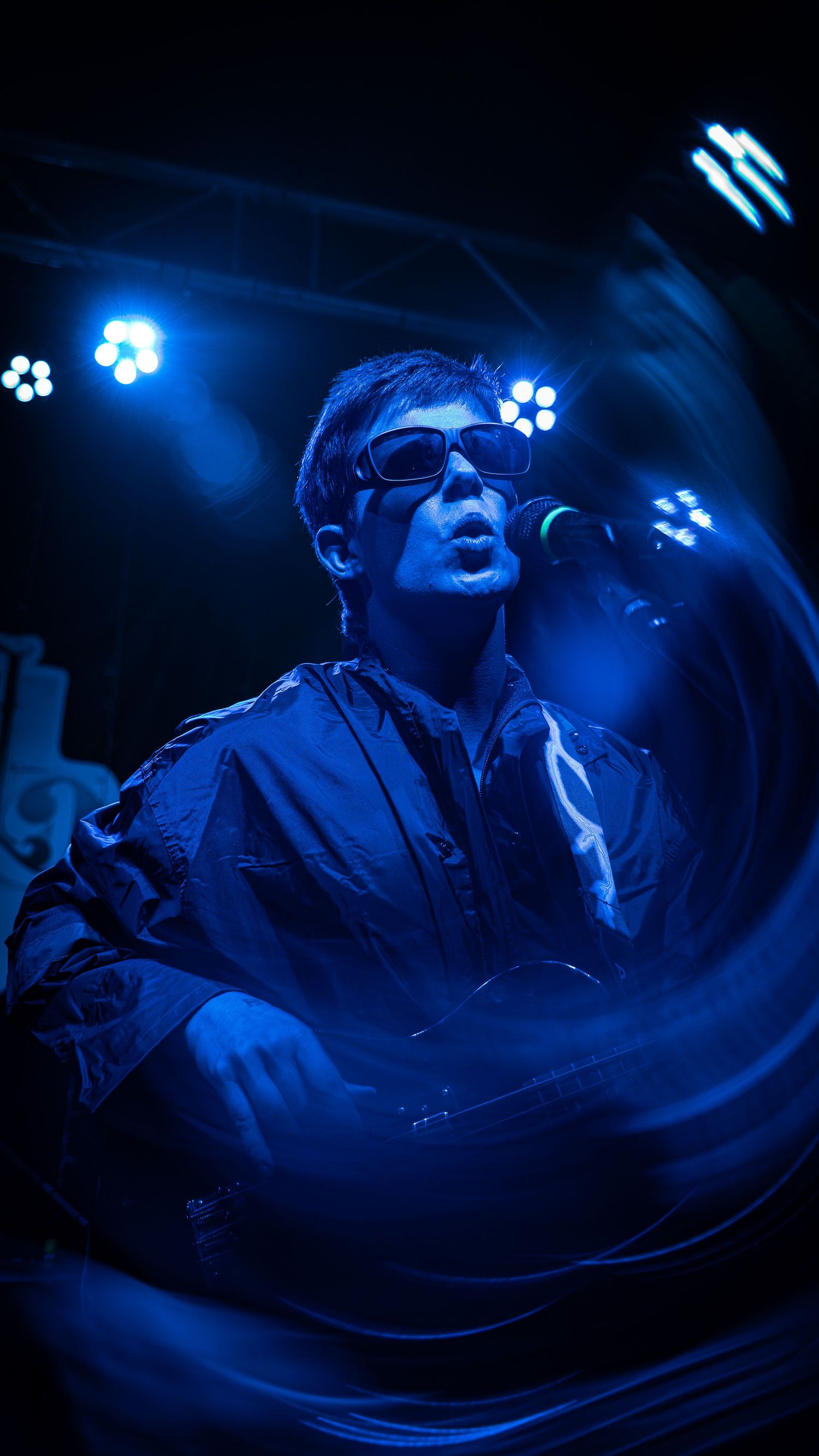 A performer in sunglasses sings on stage bathed in dramatic blue lighting during a concert performance.