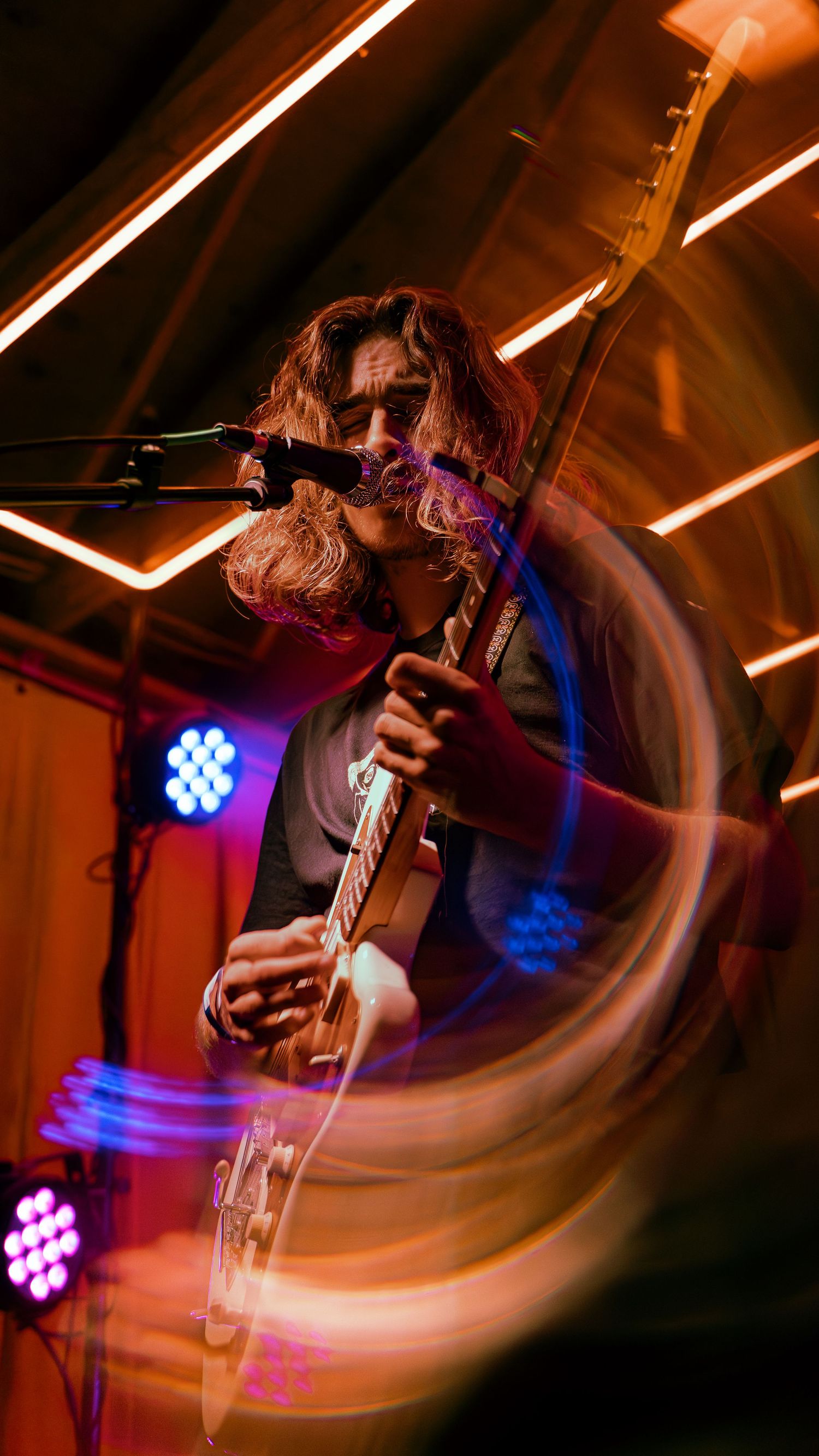 Musician performs with blurred motion effect under warm stage lighting with blue and purple LED accents in background.