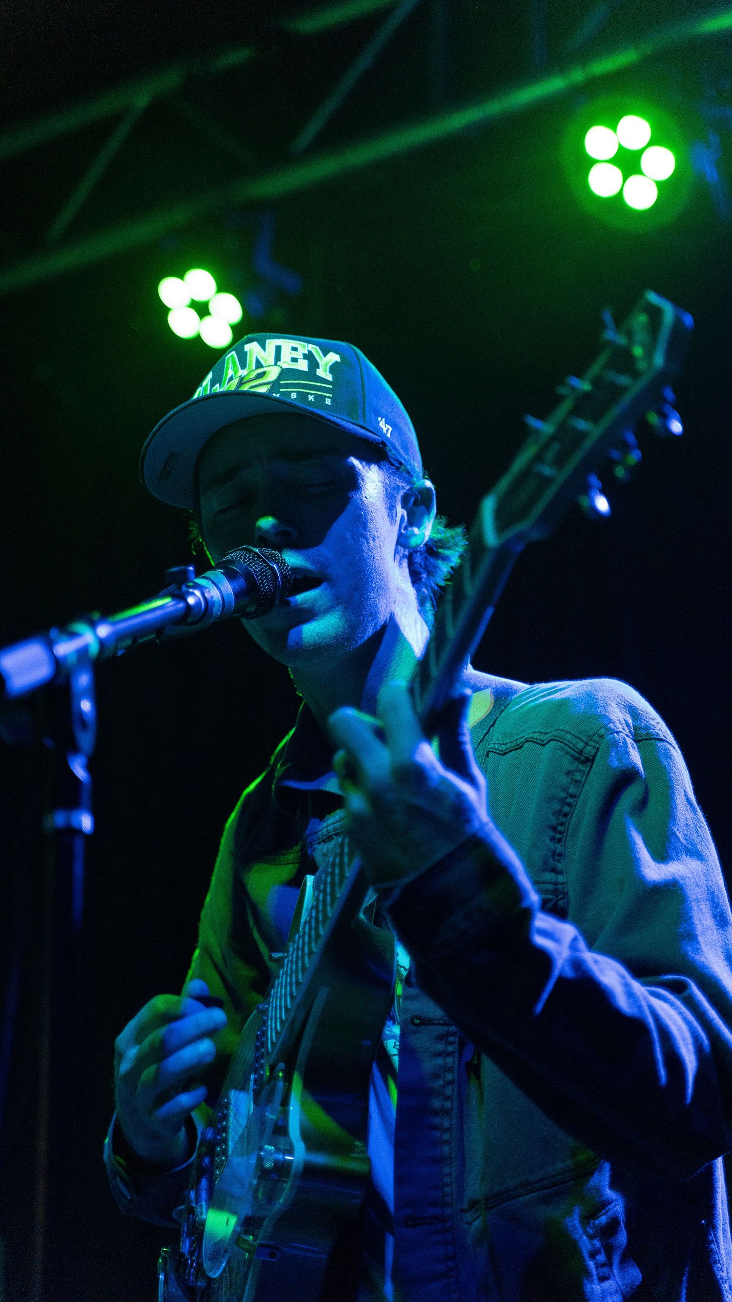 Performer in a baseball cap singing into microphone under green stage lighting.