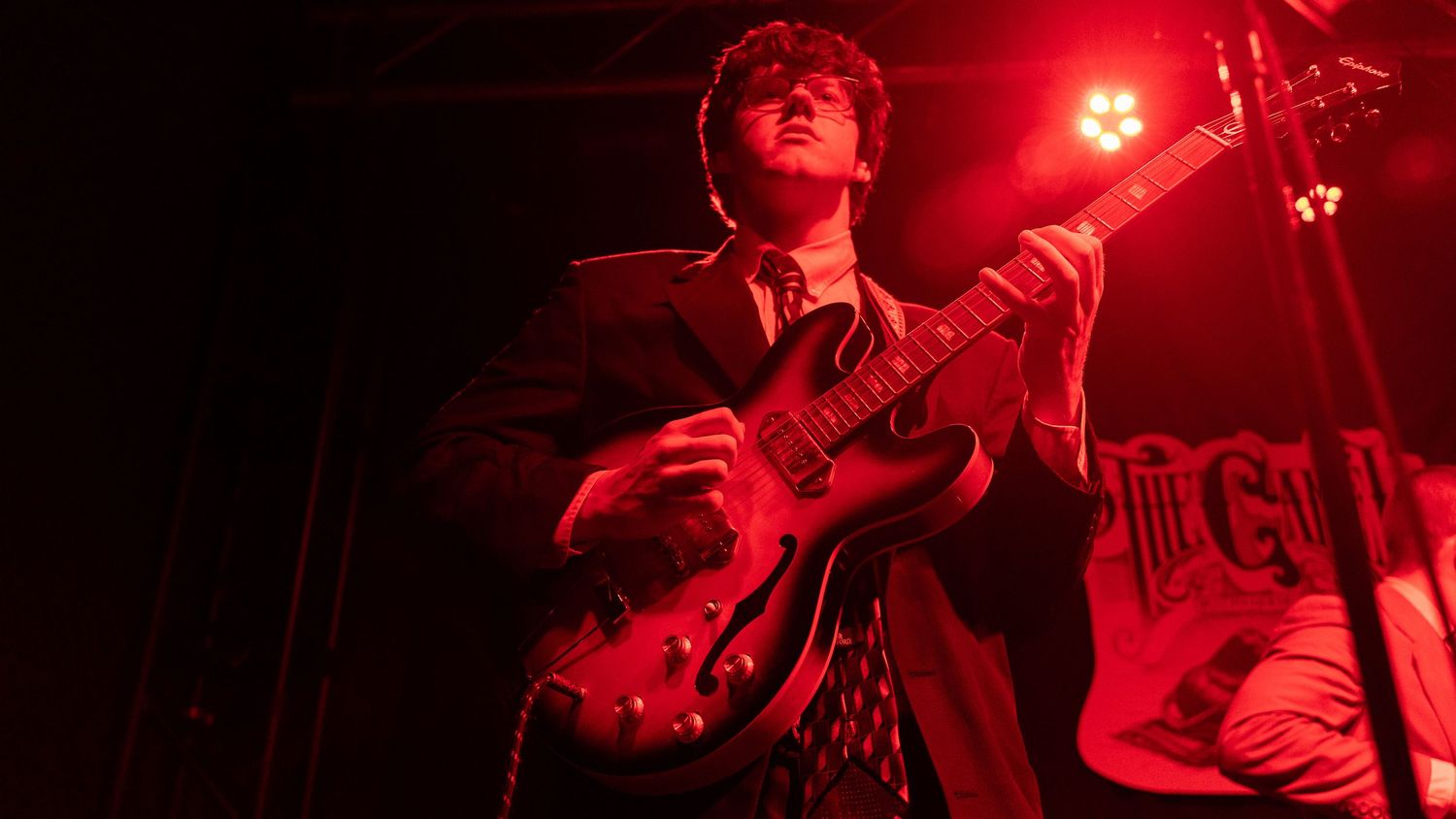 Guitarist performing on stage illuminated by intense red concert lighting.