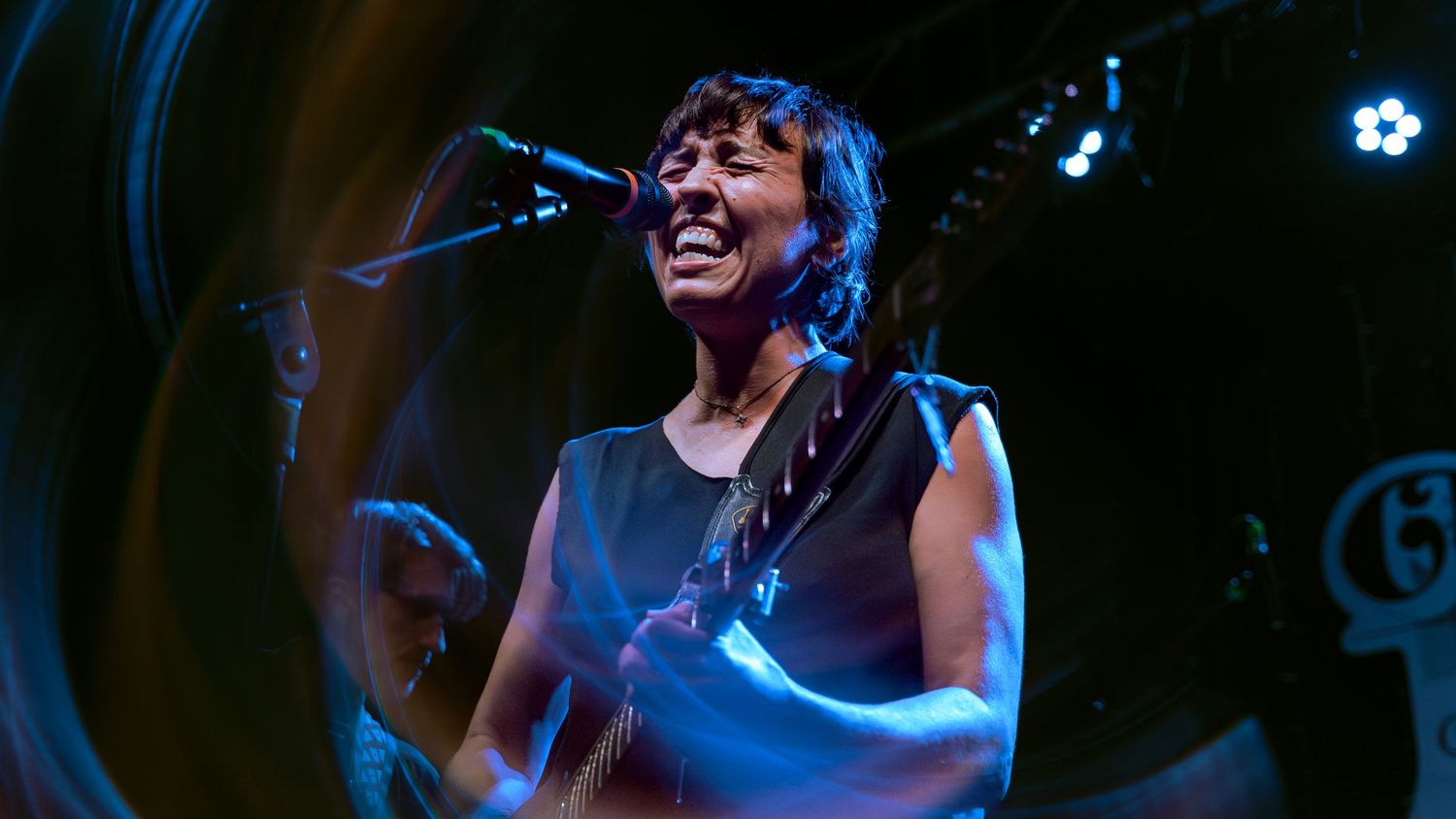 Concert performers playing music on stage with dramatic blue lighting and spotlights creating an energetic atmosphere.