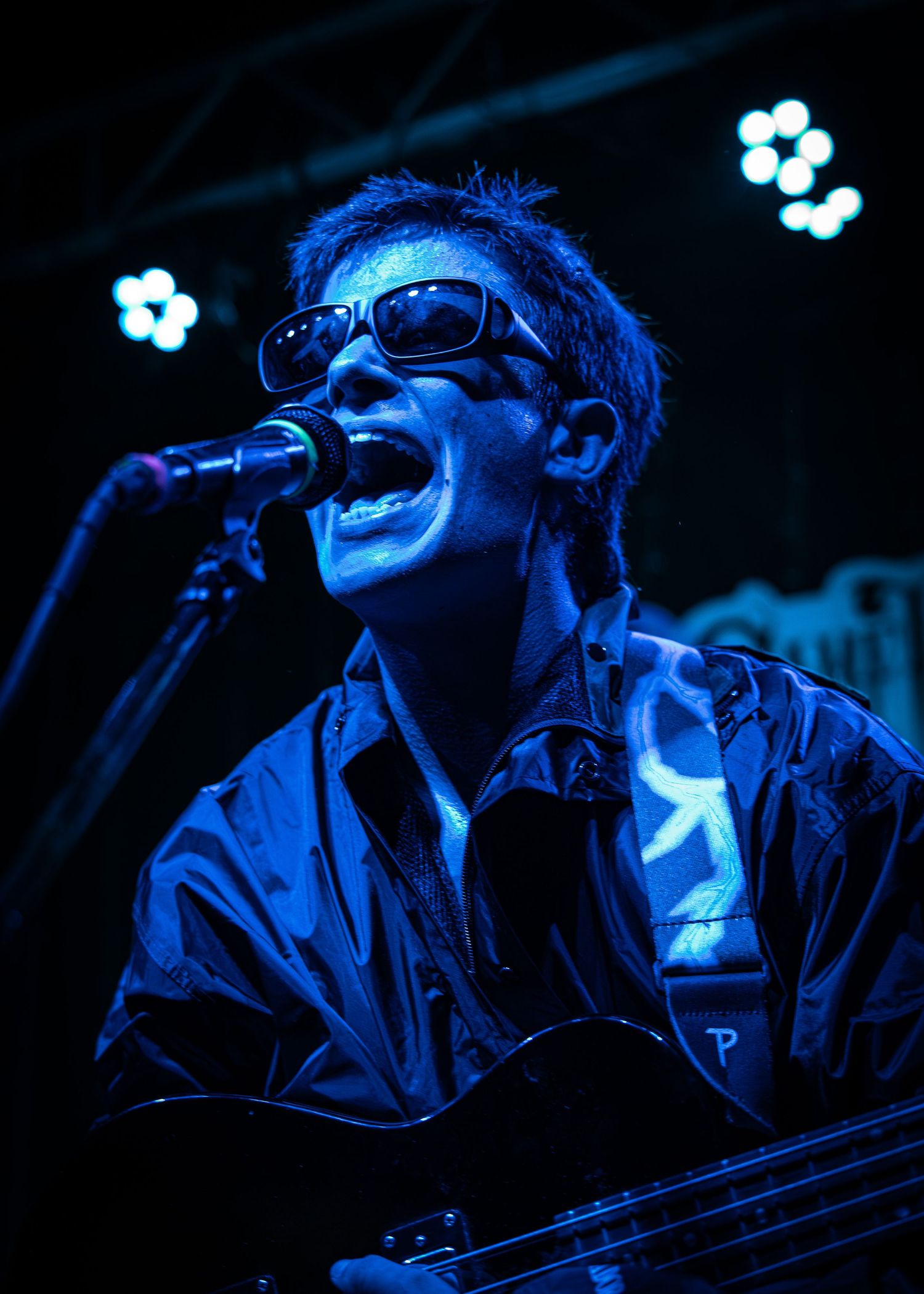 Musician performing on stage under blue lighting wearing sunglasses and jacket while singing into microphone.