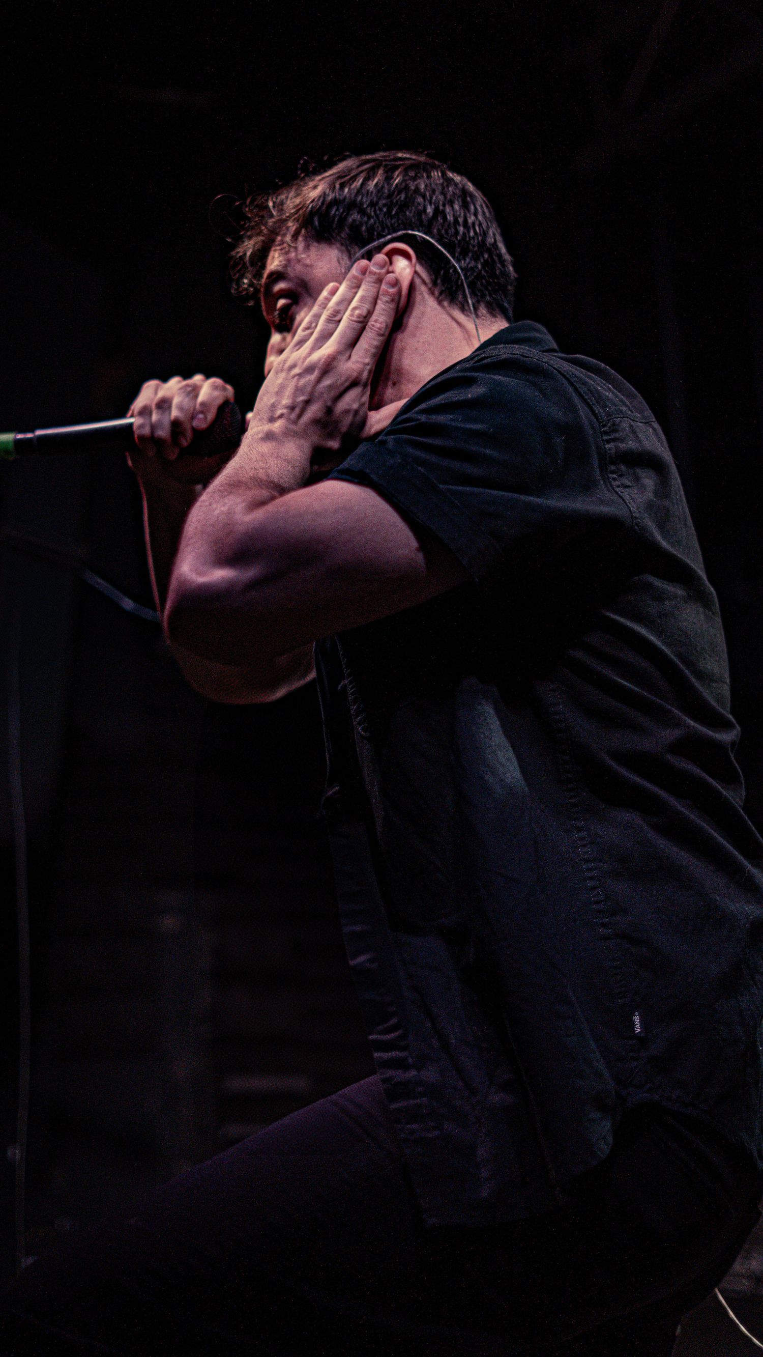 Dark silhouette of a performer in black clothing singing into a microphone on stage against dark background.