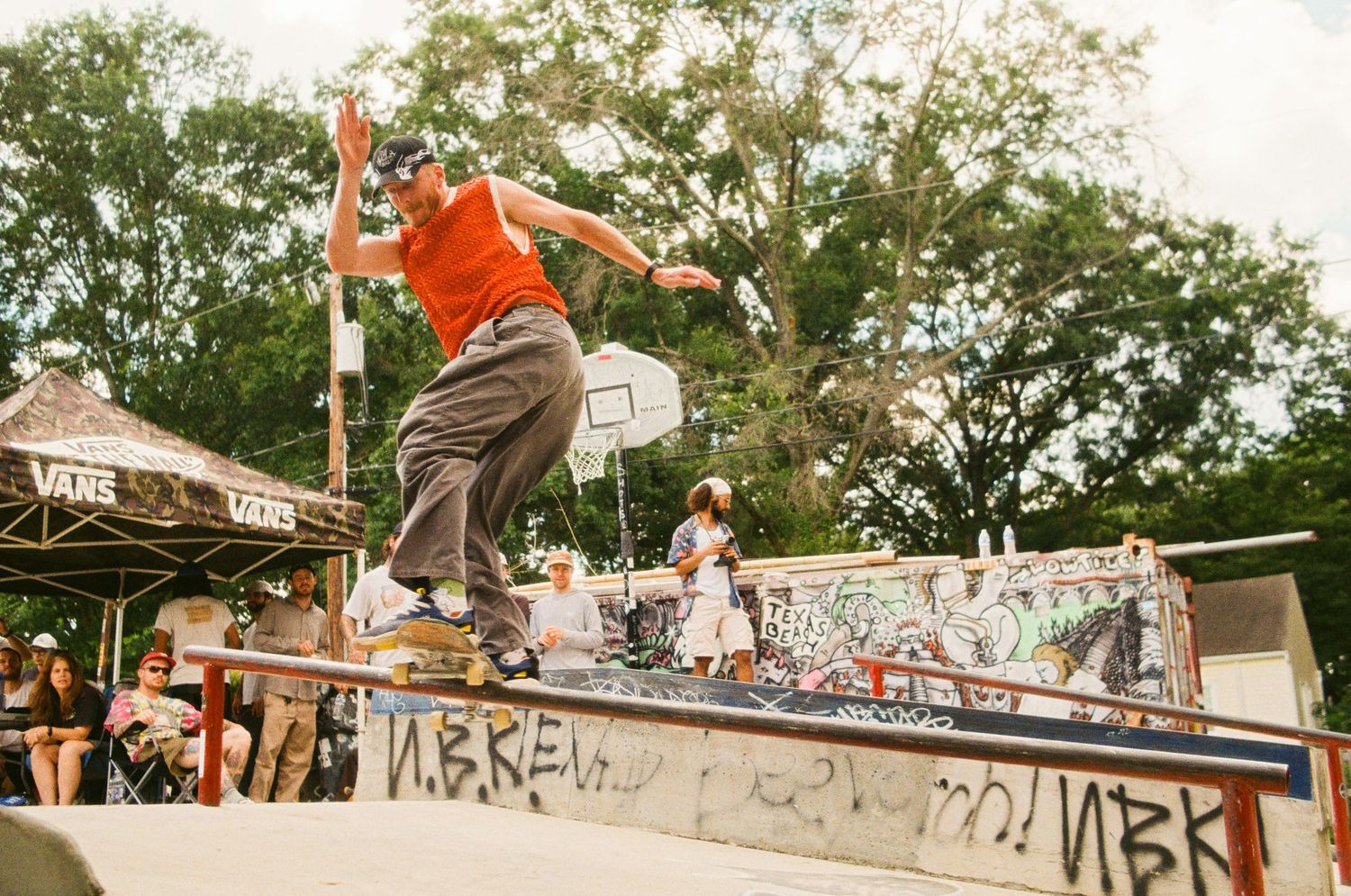 Skateboarder performs aerial maneuver on wooden ramp while others observe during outdoor skate event.