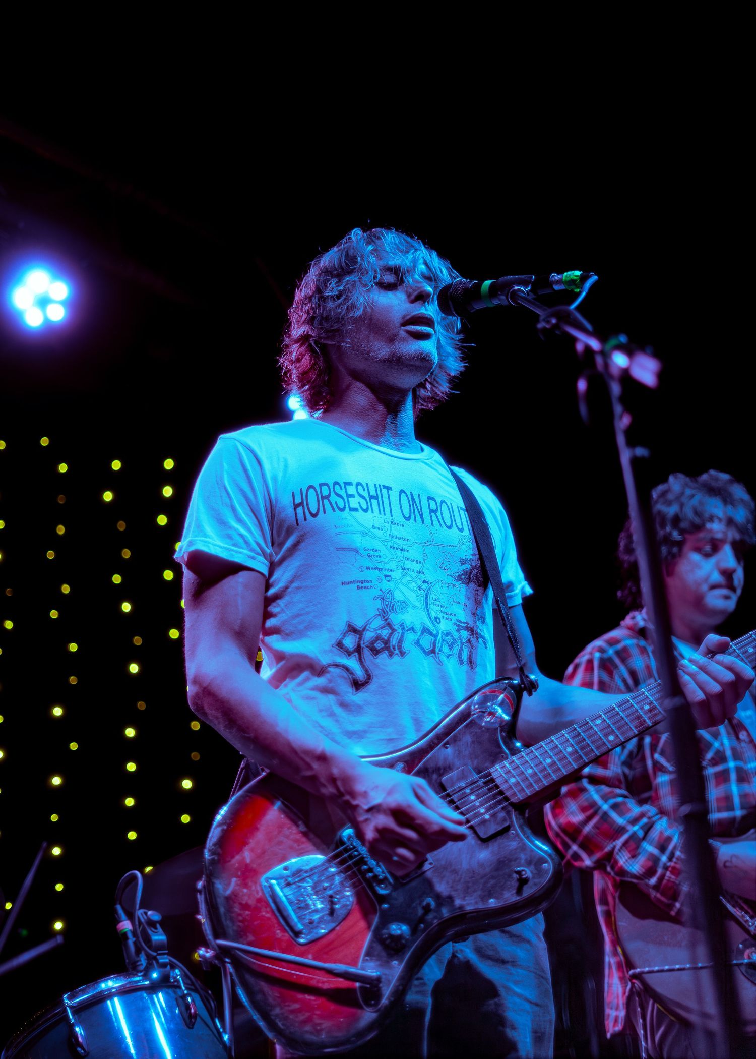 Performer in a light blue t-shirt plays electric guitar on stage under dramatic blue lighting during a concert.