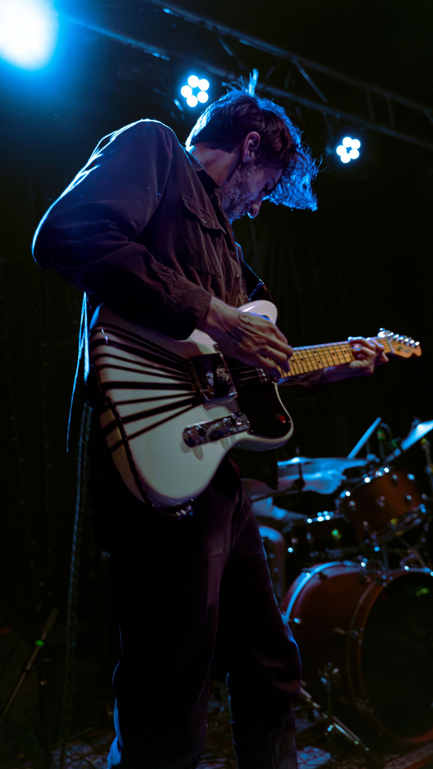 Musicians perform on a dark stage lit with blue lights while playing electric guitars in dramatic silhouettes.