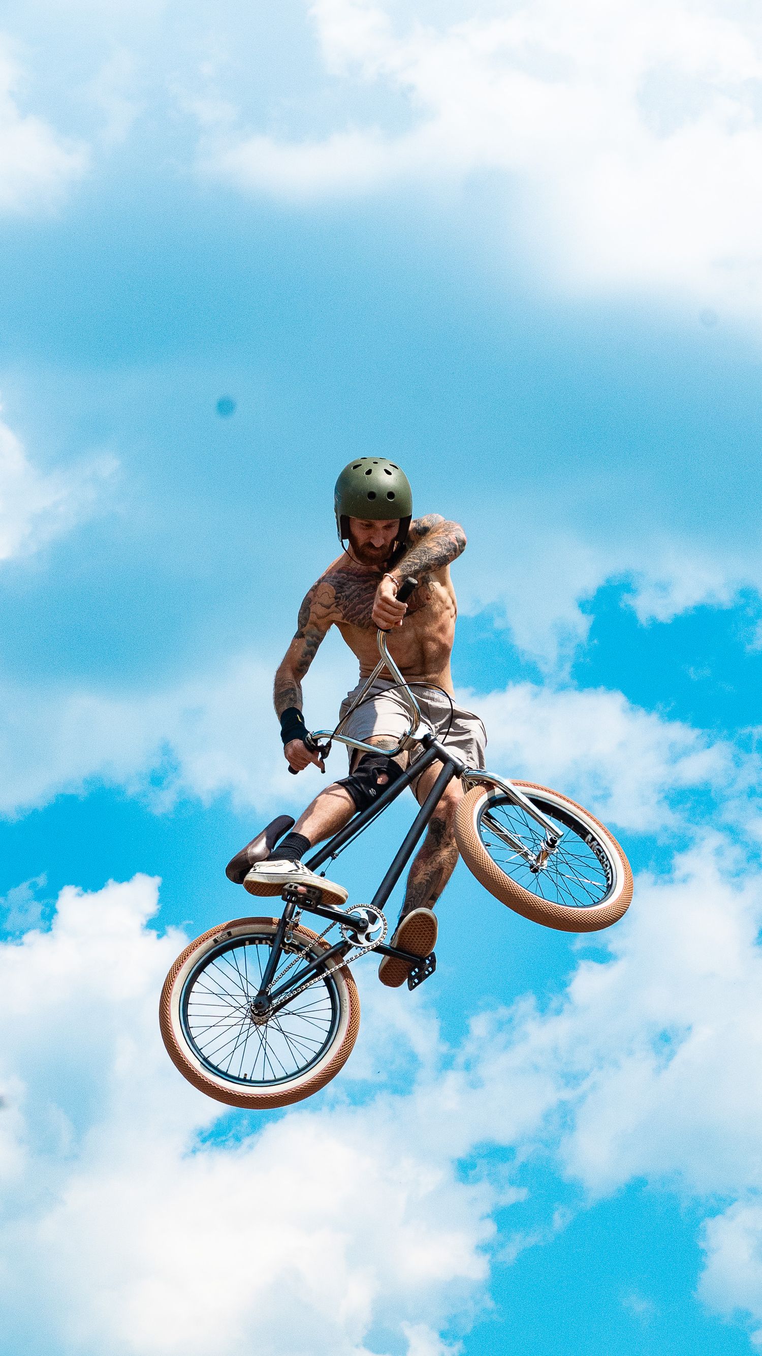 BMX rider performs airborne stunt against blue sky and white clouds while wearing protective gear.
