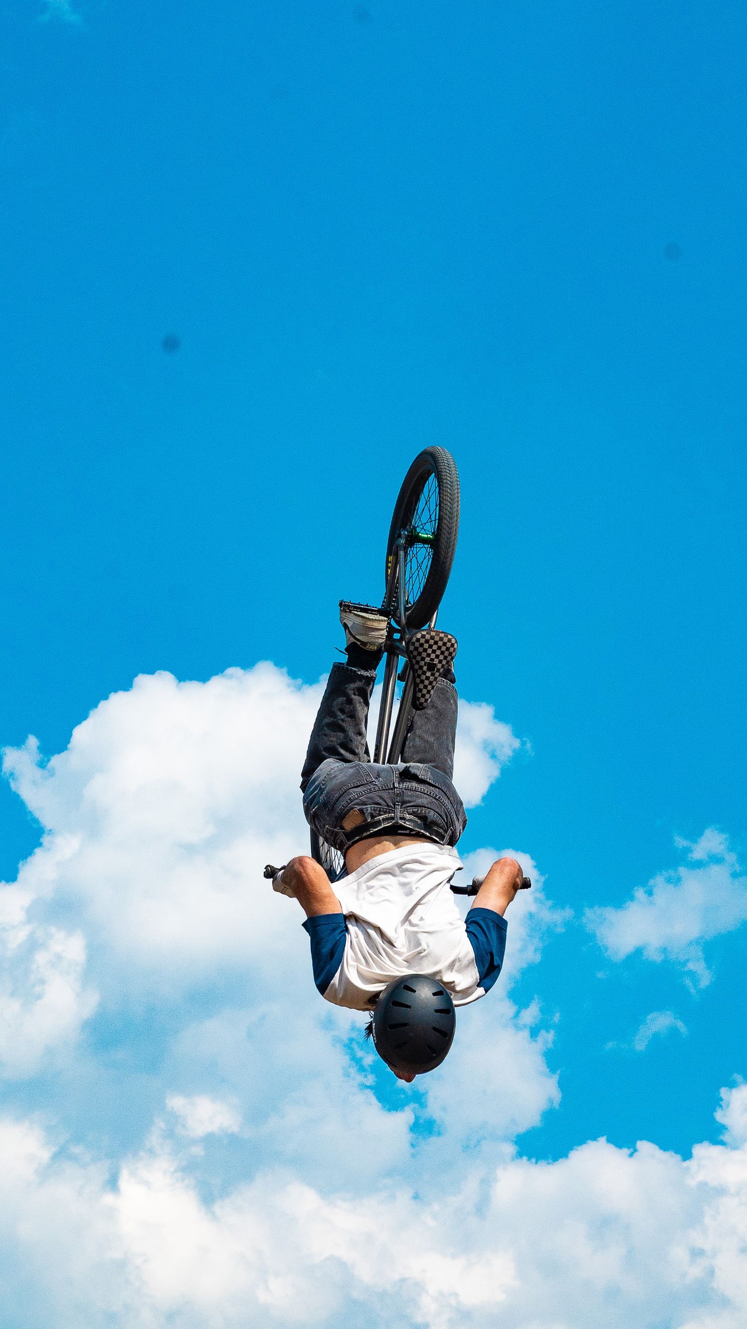 BMX rider executes an inverted aerial maneuver against blue sky and white clouds.