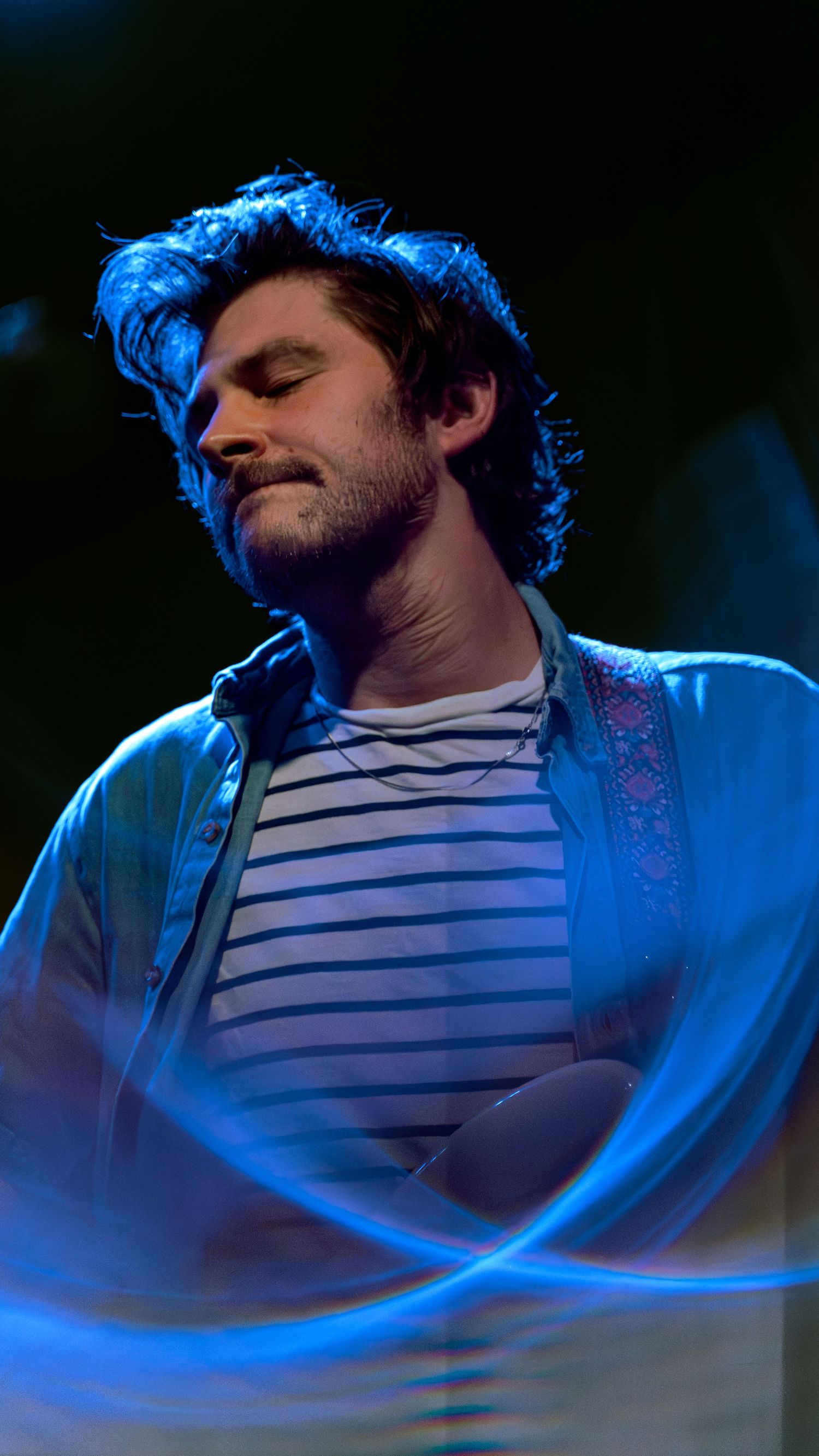 Musician in striped shirt performs passionately on stage under dramatic blue lighting during a concert performance.