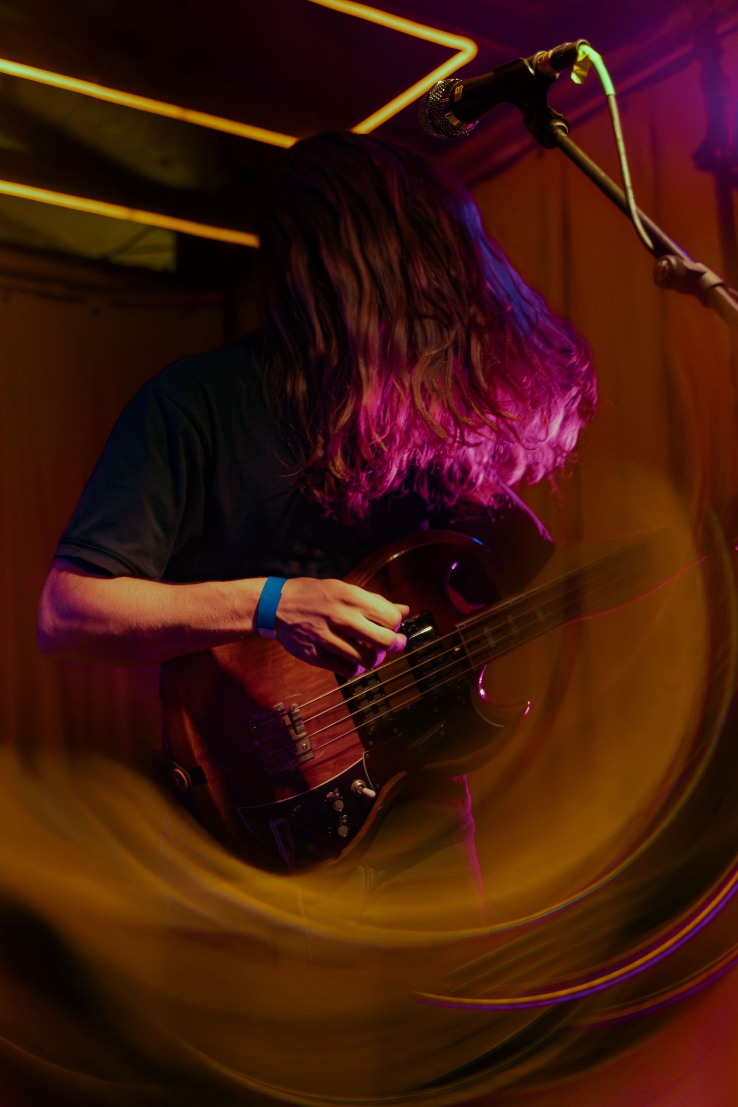 Musician with long dark hair plays bass guitar under colorful purple and yellow stage lighting.
