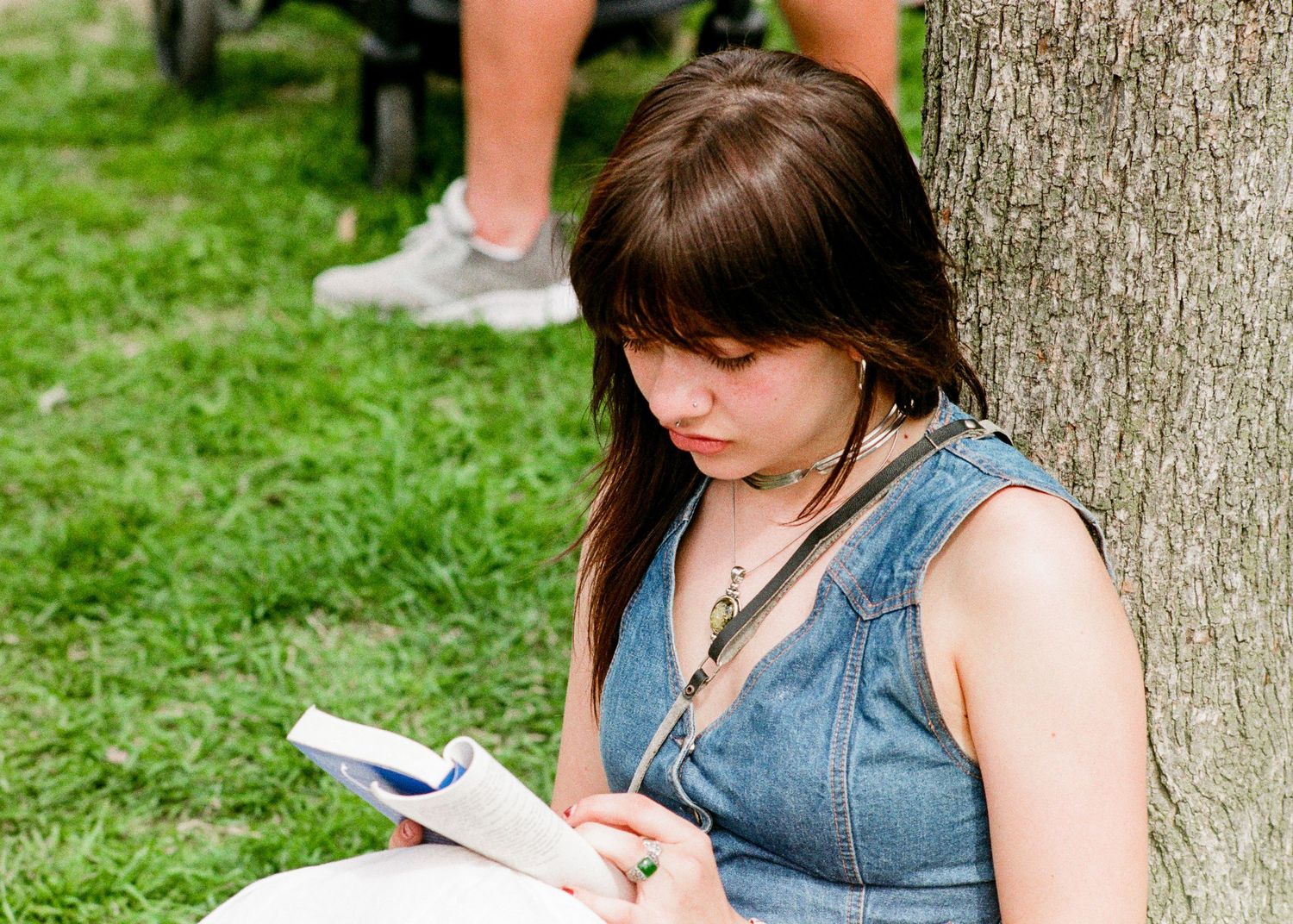 Someone in a denim top sits against a tree trunk while reading on a grassy lawn outdoors.