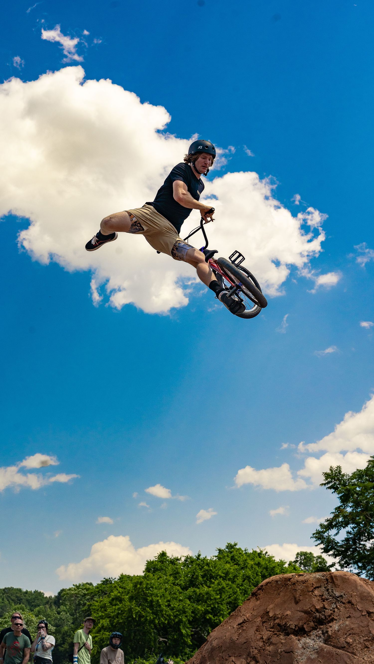 BMX rider soars through air off dirt jump with spectators watching below.