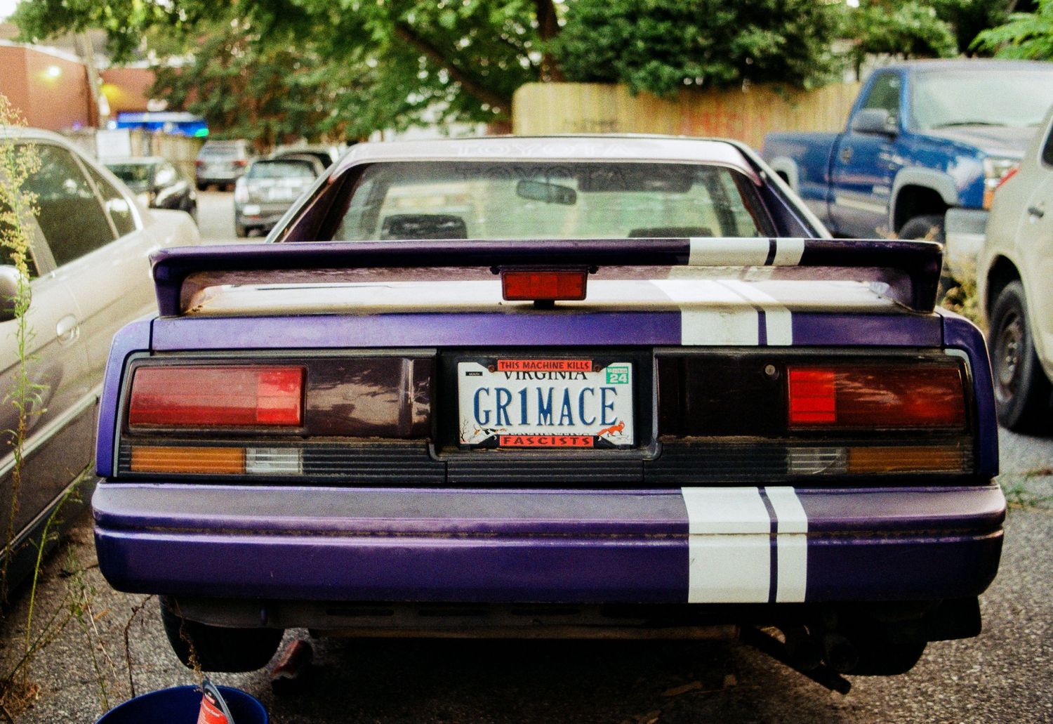 Purple vintage car with racing stripes shown from rear view in parking lot.