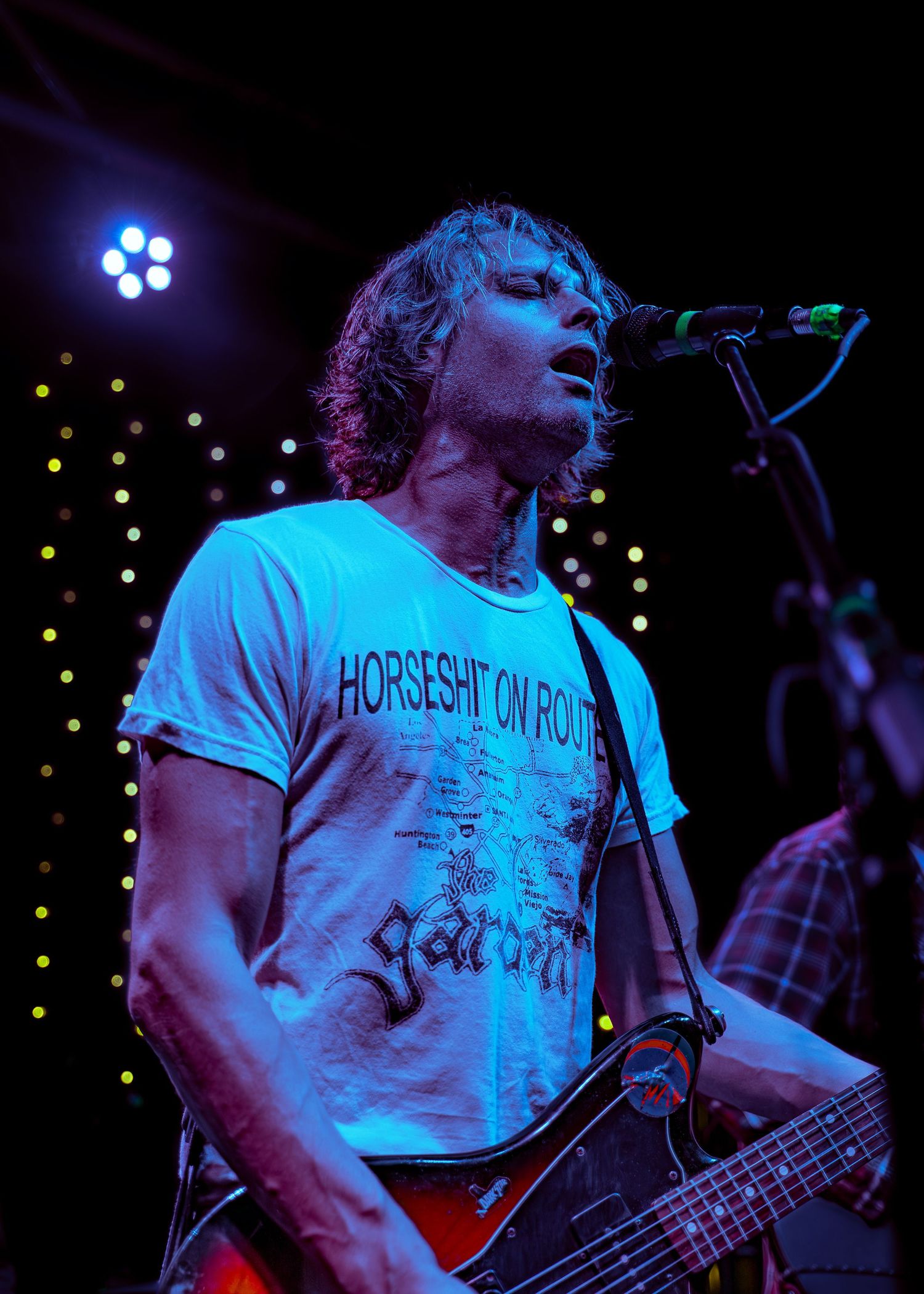 A musician performs on stage illuminated by blue lighting while wearing a white t-shirt and playing a bass guitar.