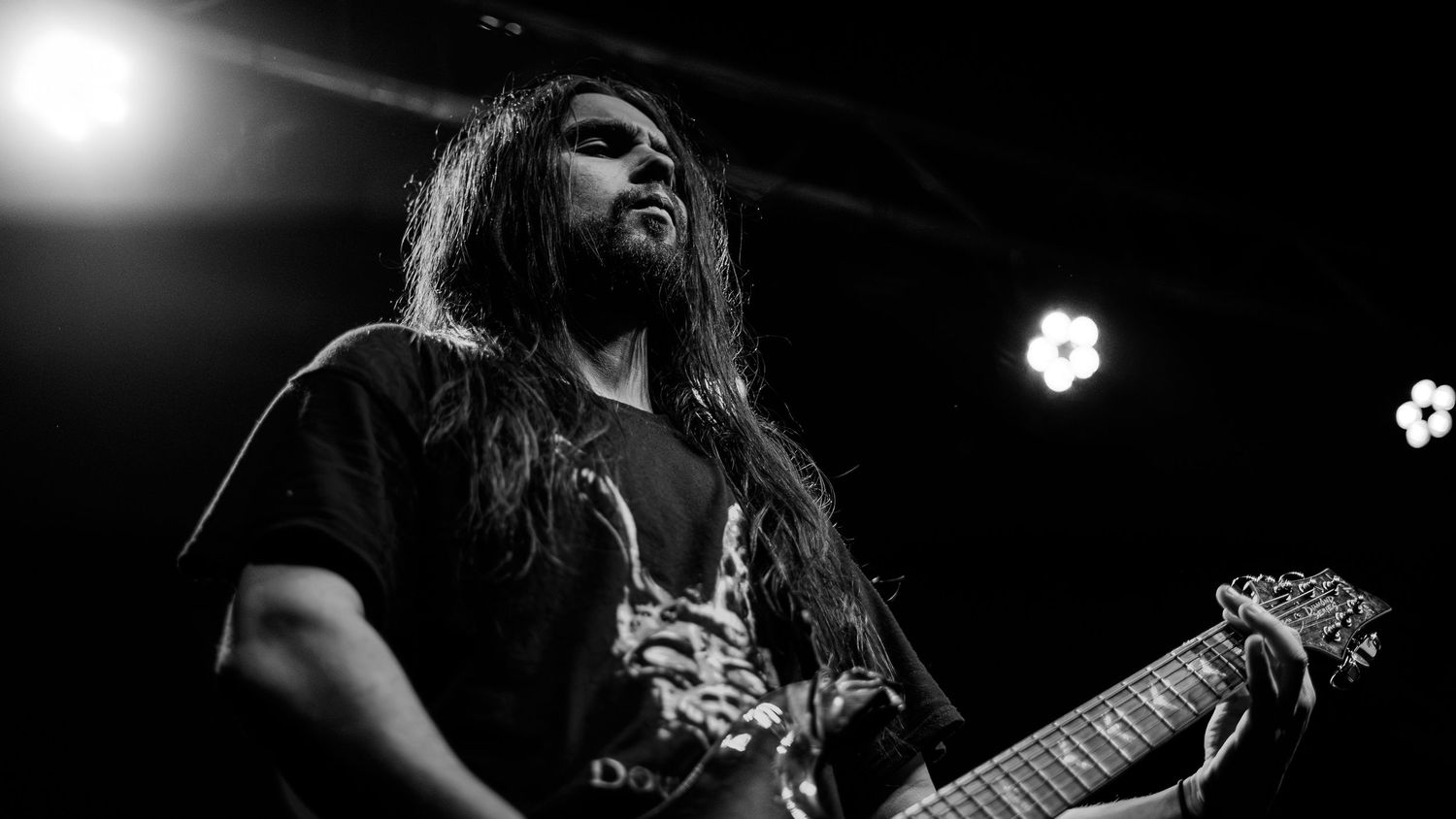 Black and white photo of a long-haired guitarist performing on stage with bright spotlights in the background.