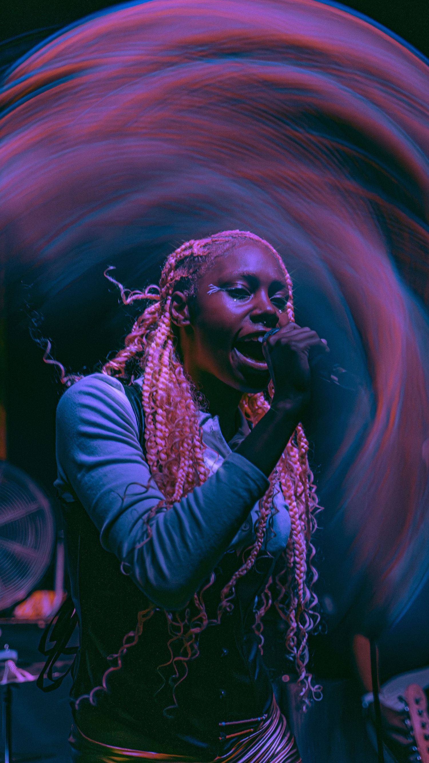 A performer singing passionately on stage with long braids, illuminated by dramatic purple and blue lighting effects.