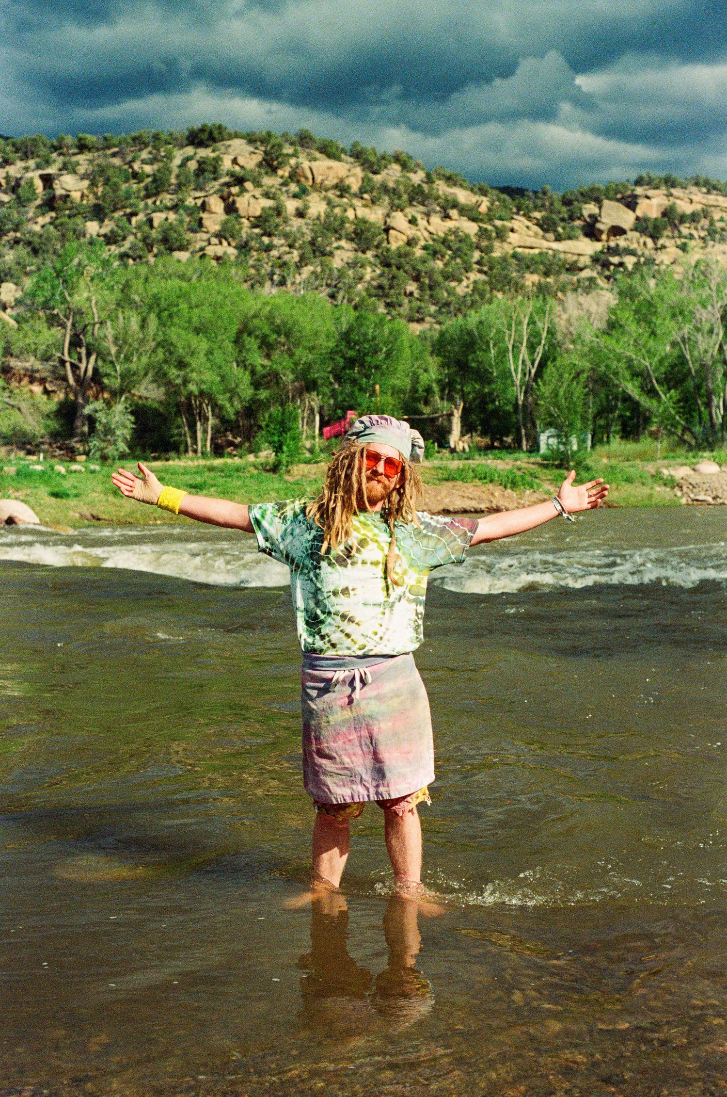 Person standing joyfully in shallow river water with arms spread wide against desert cliffs.