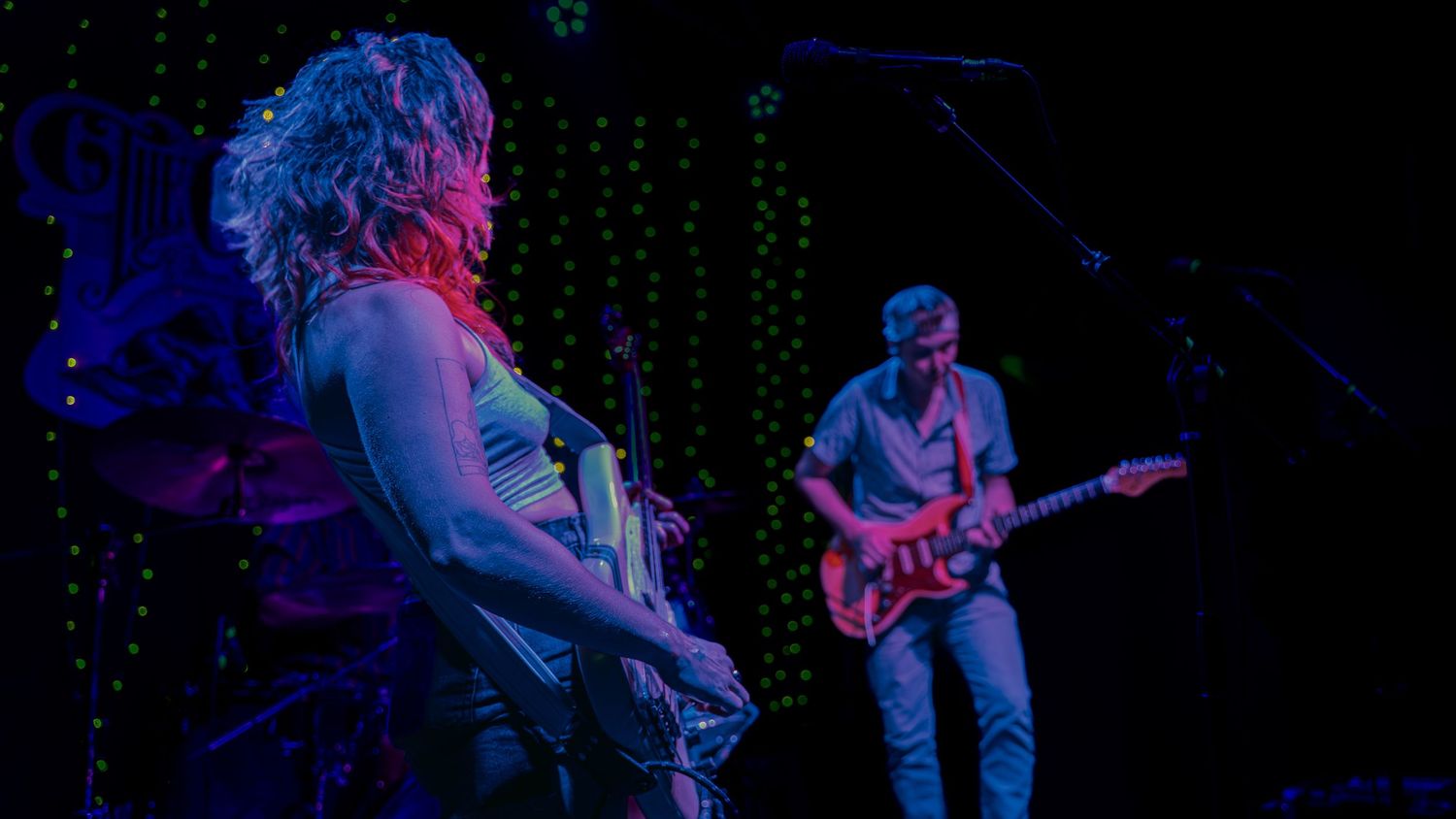 Musicians perform on a dark stage illuminated by blue and pink lighting with a guitarist visible in the background.