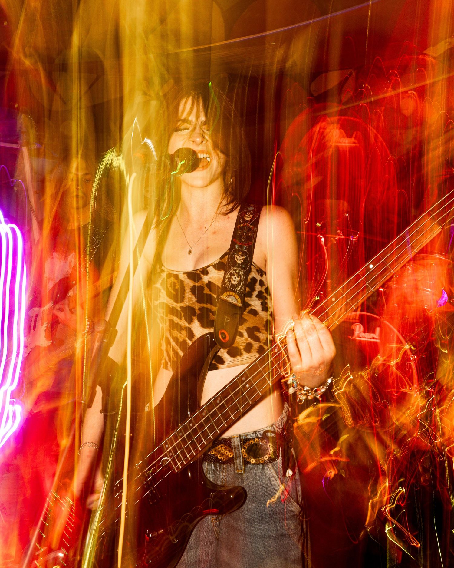 Concert performer in leopard print top playing bass guitar under colorful stage lighting effects.