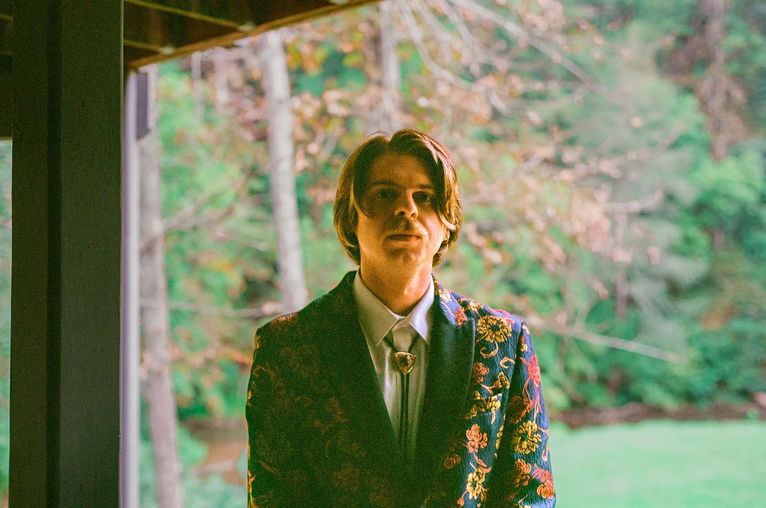 Formal portrait of someone in a suit and tie standing by a window with greenery in the background.