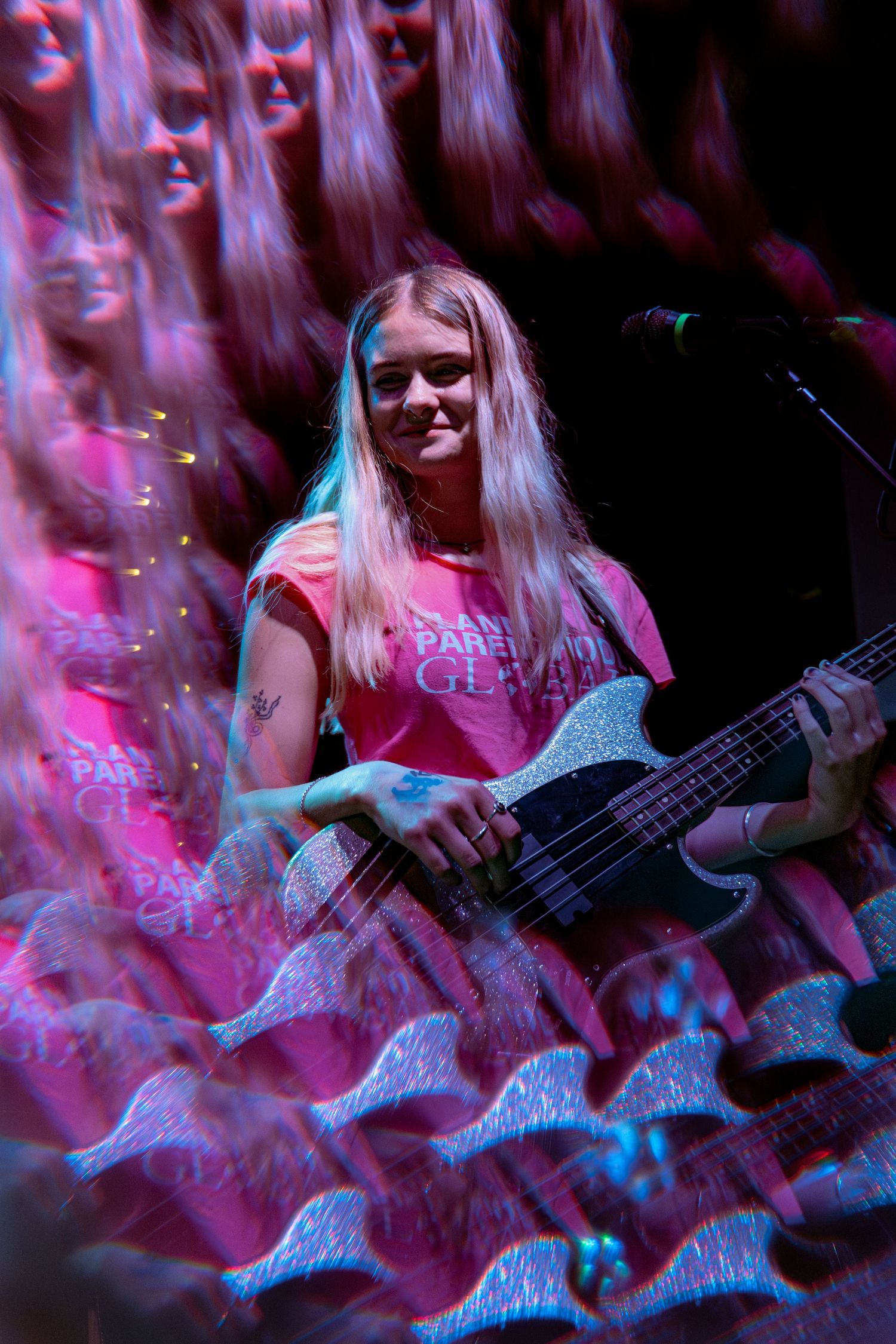 A bass guitarist performs on stage bathed in dramatic pink and blue lighting effects during a live concert performance.