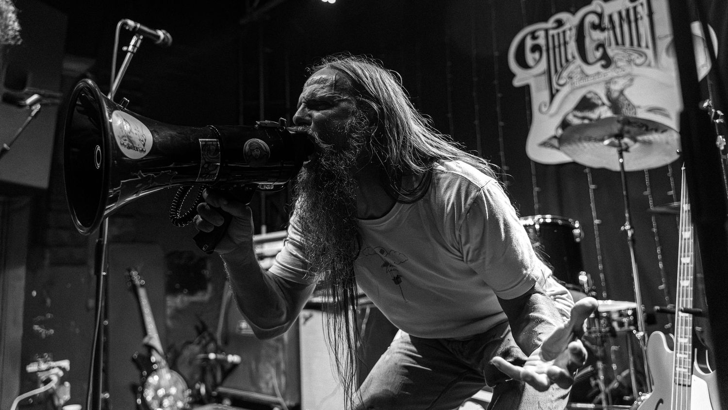 A singer performs passionately on stage with long hair and glasses at a rock concert with band equipment visible in background.