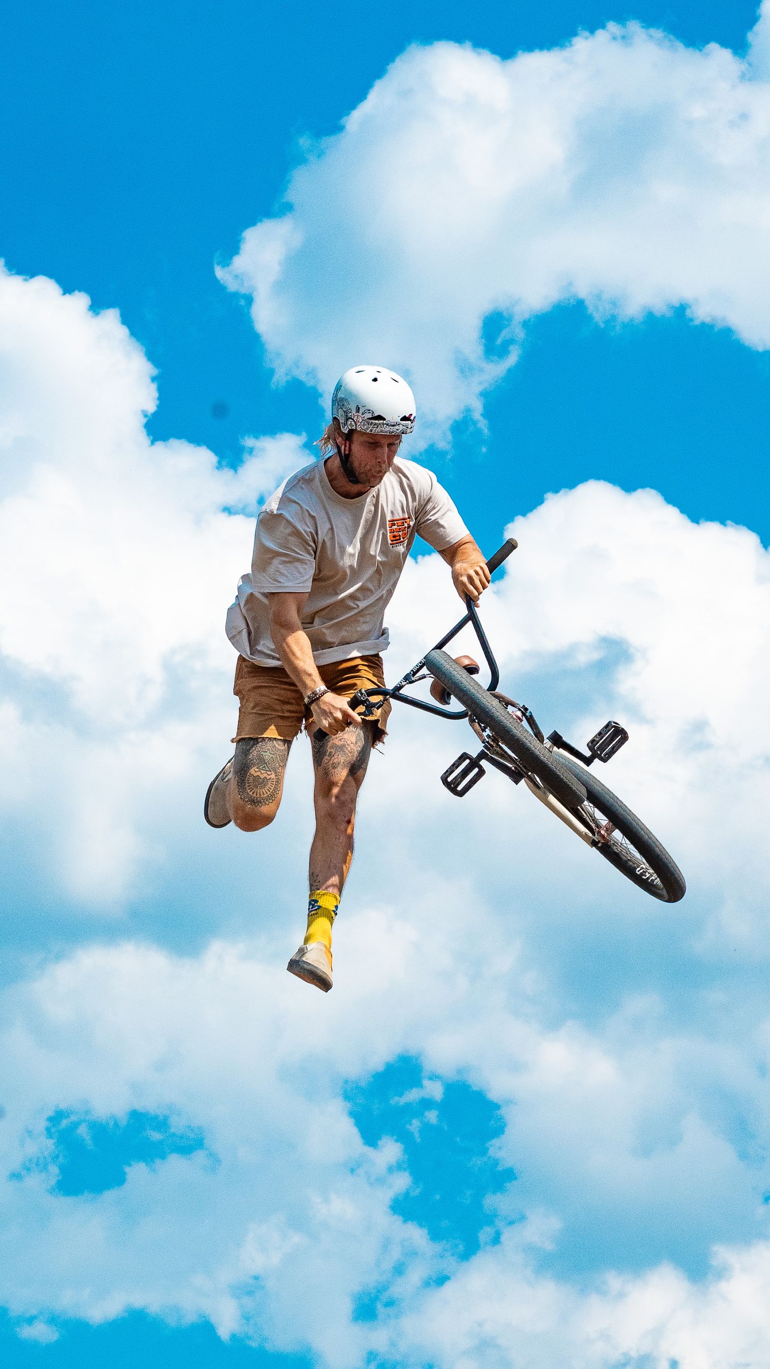 Rider performs no-handed jump trick on BMX bike against puffy white clouds.