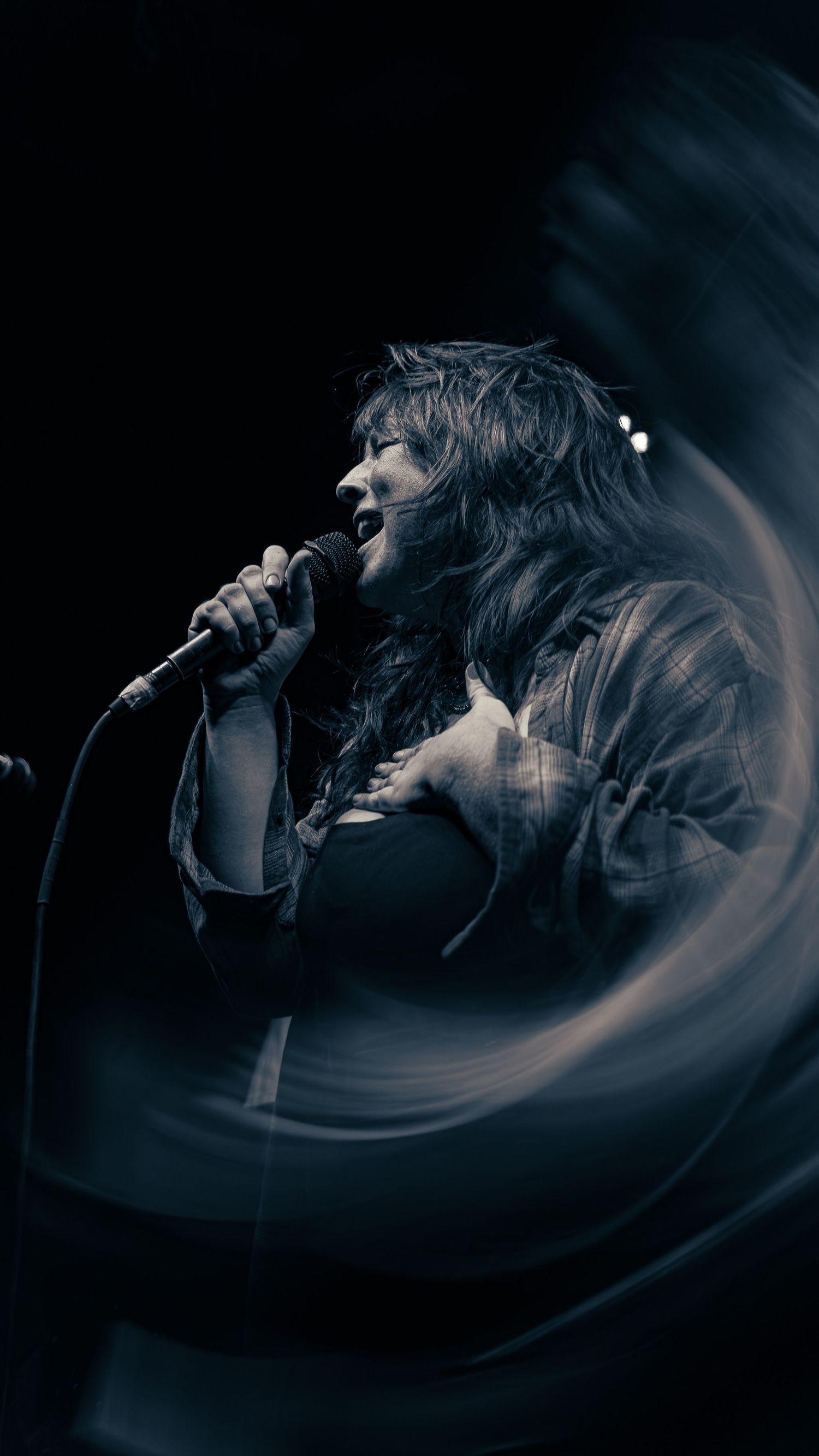 A performer sings passionately into a microphone on a dark stage with dramatic blue lighting and swirling effects.