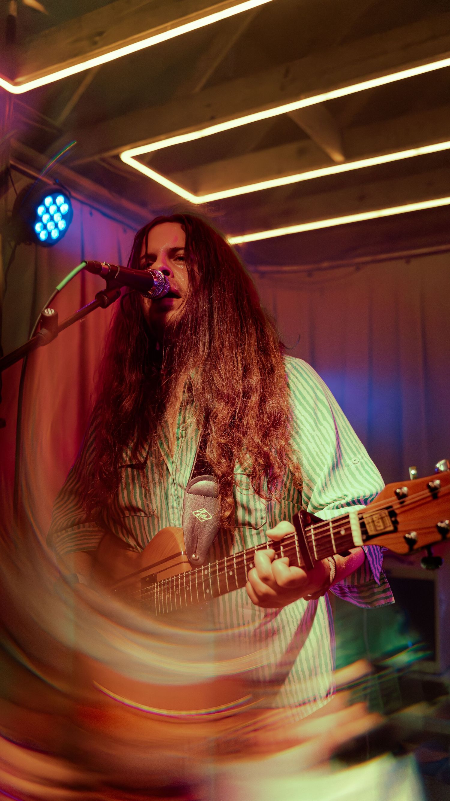 Musician with long dark hair performs on guitar under colorful stage lighting with motion blur effects.