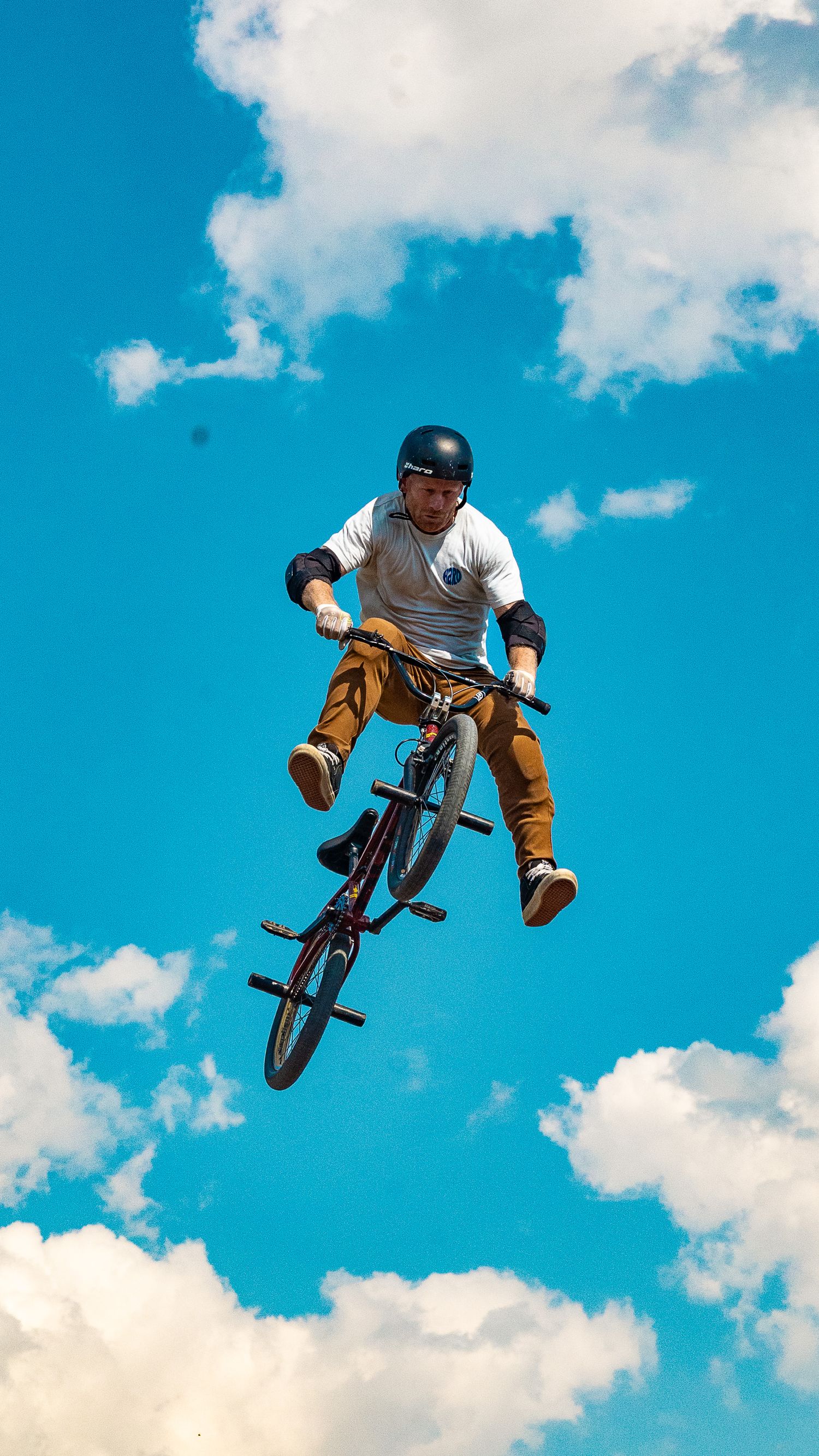 BMX rider in white shirt and orange pants performs aerial stunt against bright blue sky.
