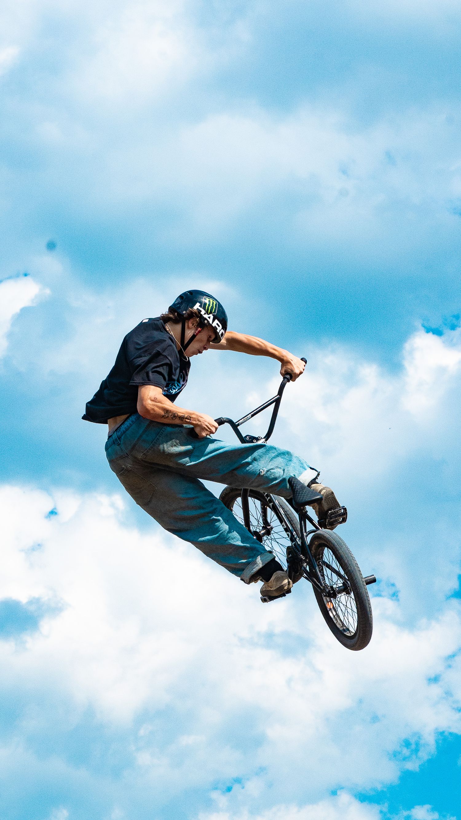 Extreme sports rider executes BMX stunt jump with blue sky and clouds in background.