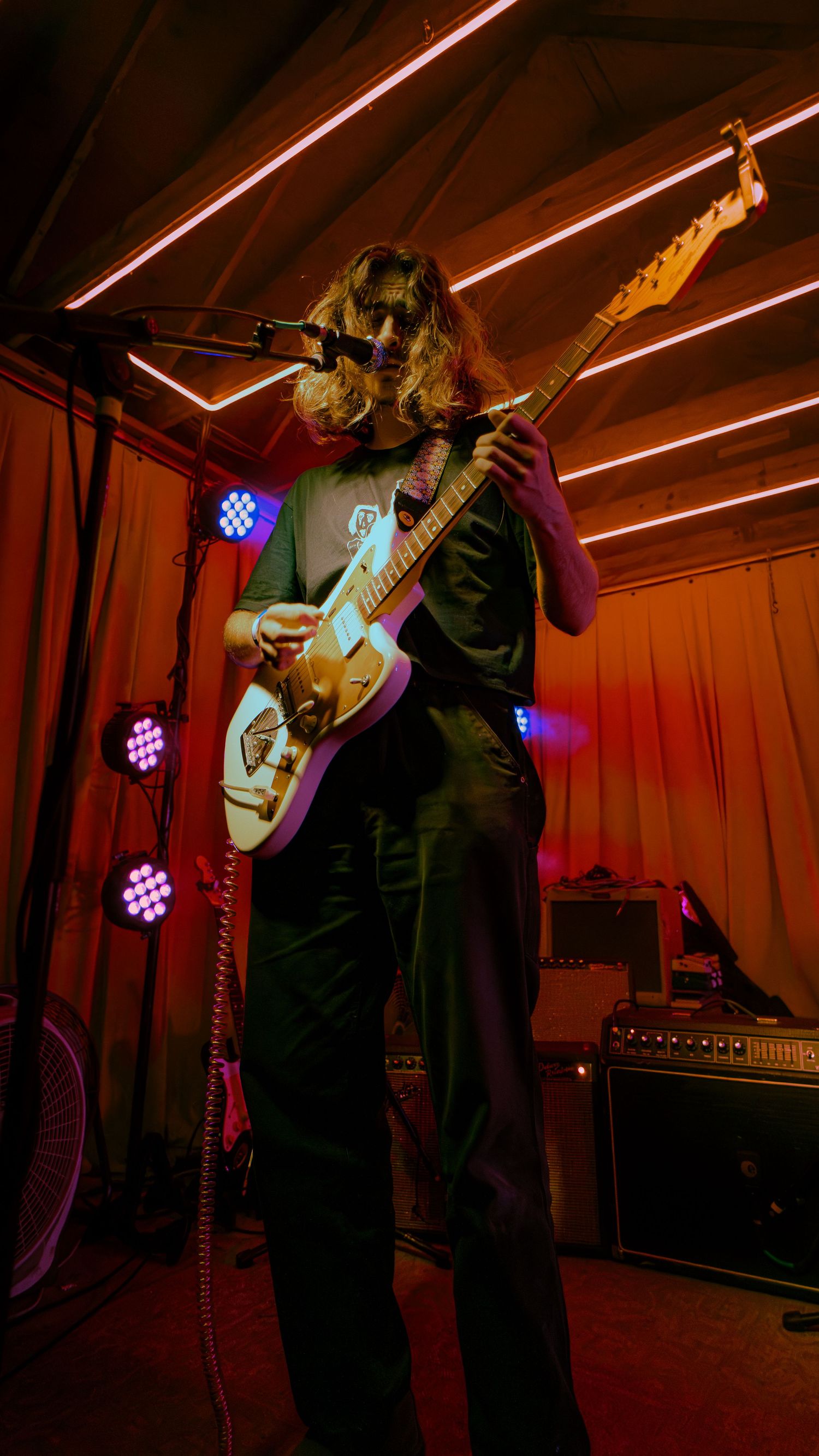 Musician performs with a guitar under atmospheric red and blue stage lighting in a dimly lit venue.
