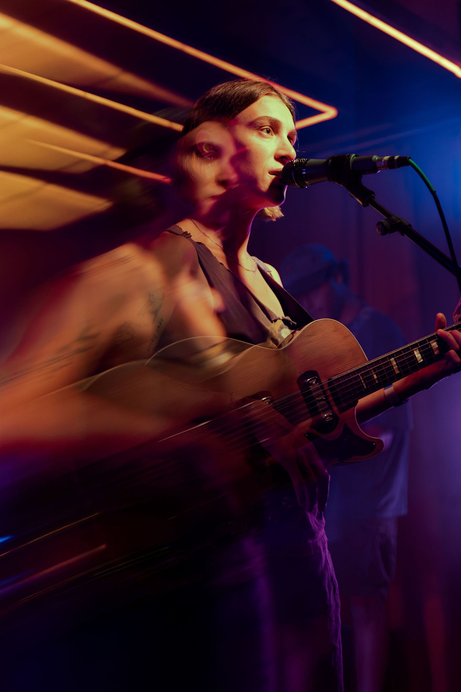 A musician performs on stage in moody blue and purple lighting while playing guitar and singing into a microphone.