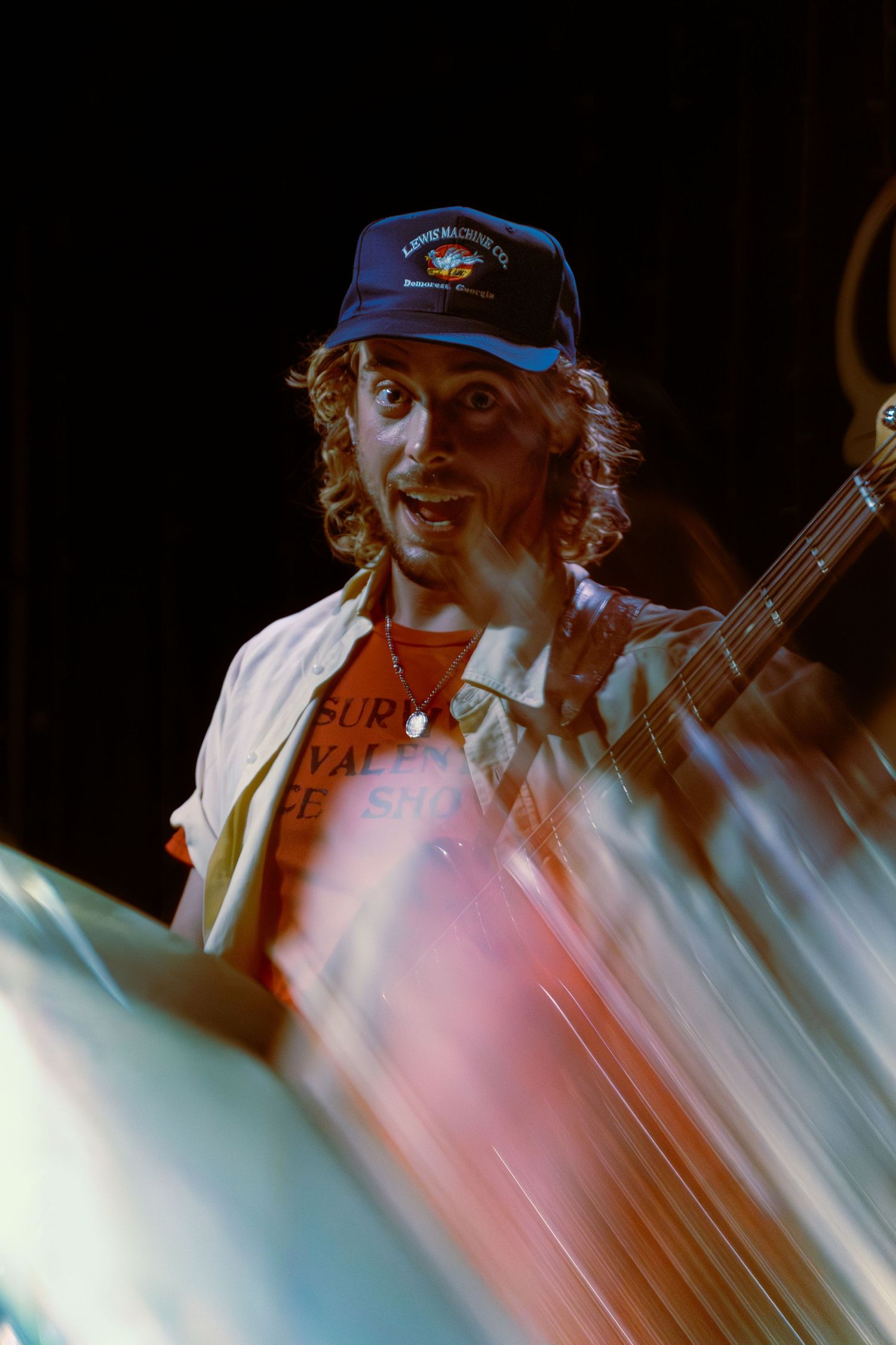 Concert performer in blue cap plays acoustic guitar on stage with dramatic lighting effects in pink and blue tones.