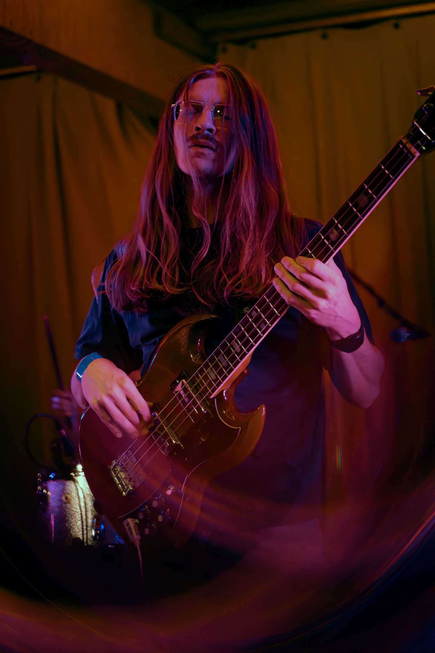Bass guitarist with long hair performs on stage in moody red lighting creating a dynamic motion blur effect during performance.