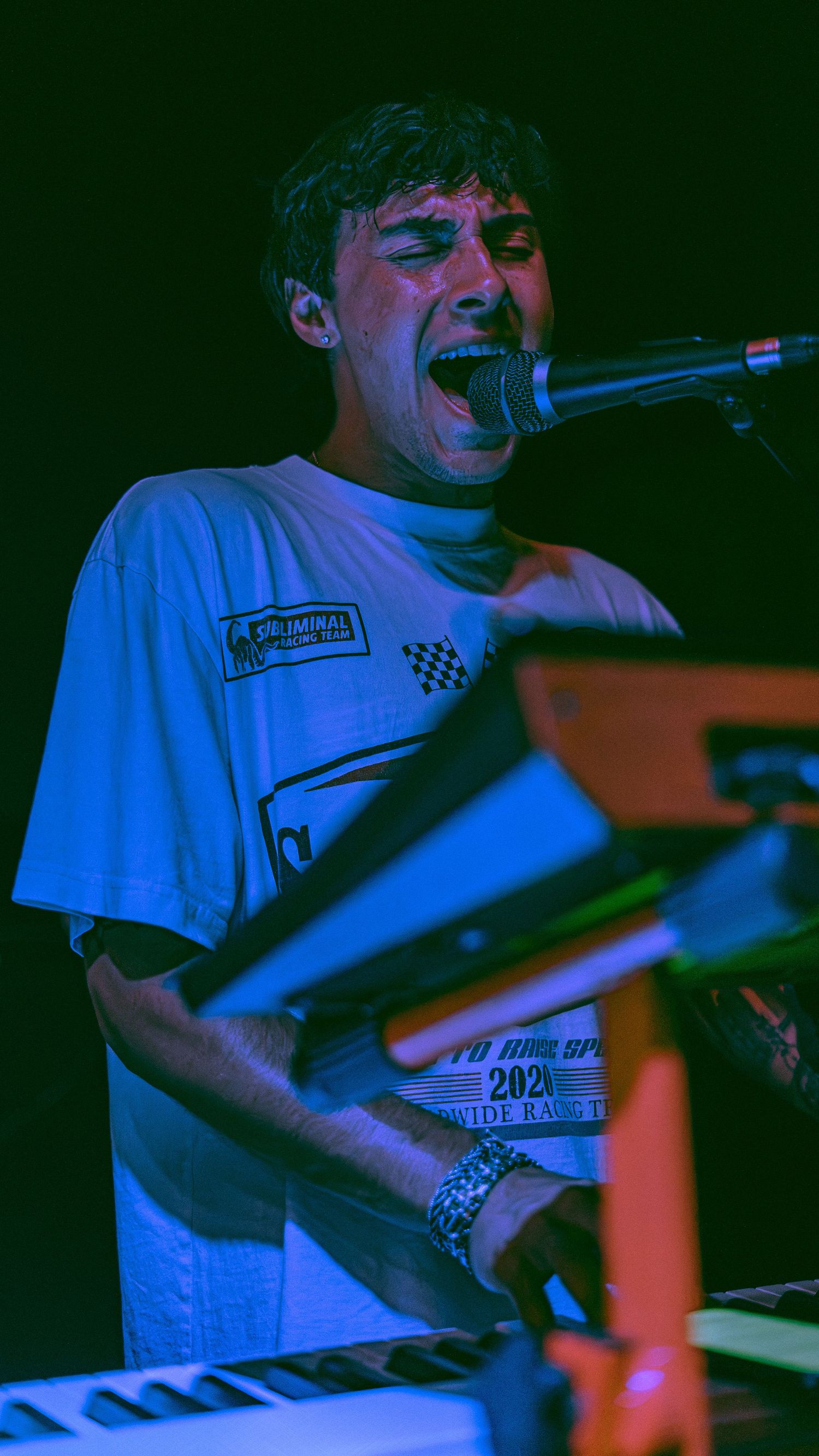 Musician performs energetically on keyboard under dramatic blue stage lighting wearing a white t-shirt.
