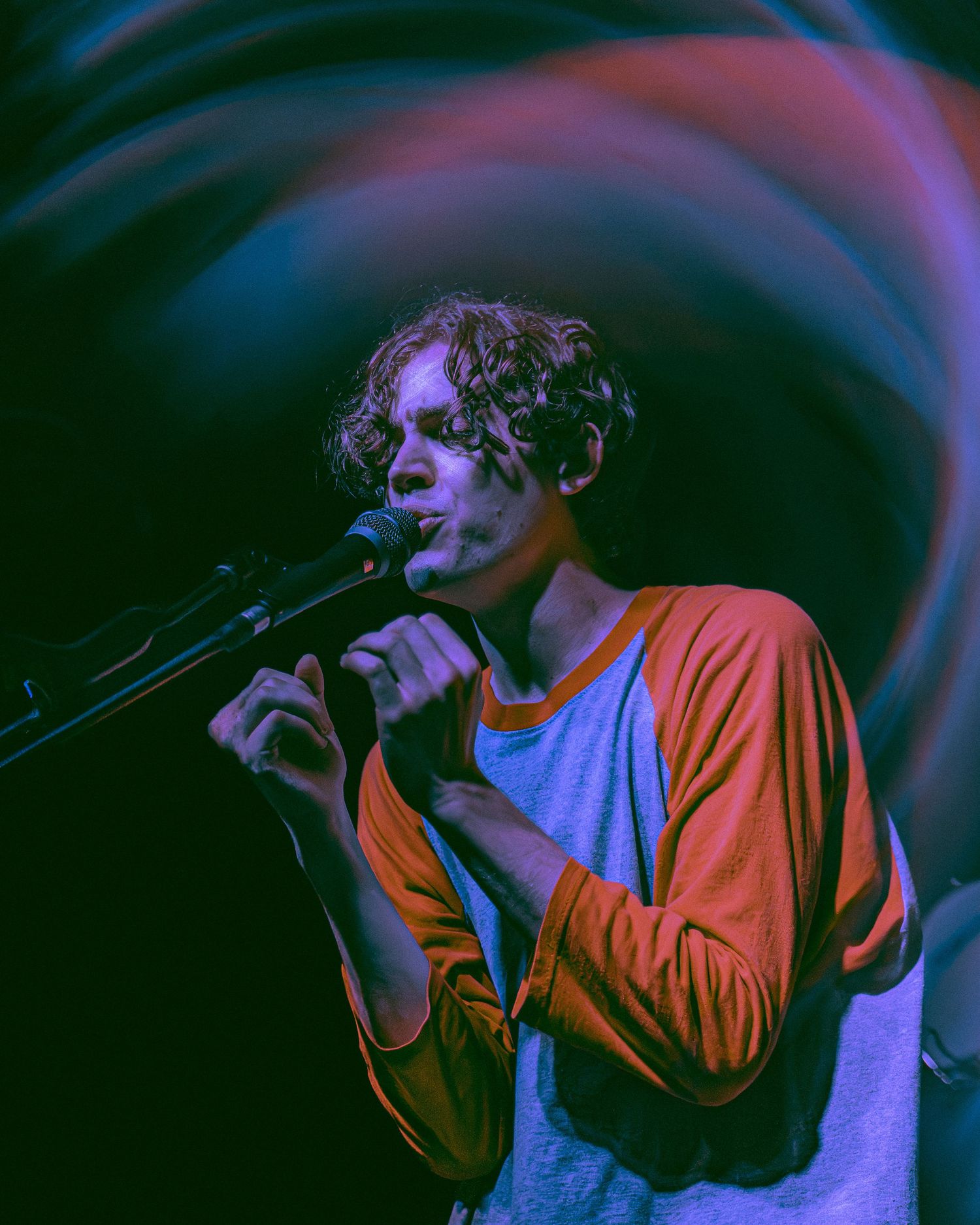 A performer in an orange and blue shirt sings into a microphone on stage with swirling purple lighting effects.