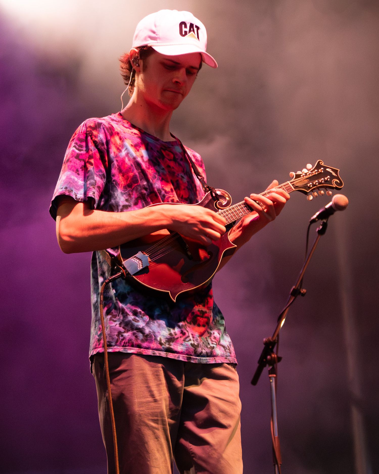 Musician in tie-dye shirt and CAP hat plays mandolin on stage during performance.