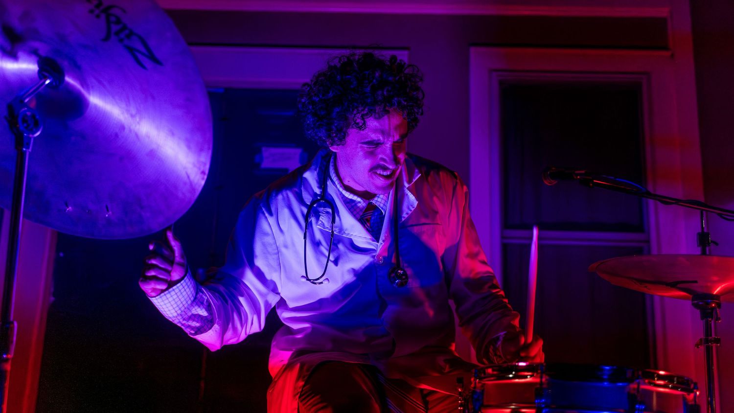Drummer performing on stage with purple stage lighting and dramatic shadows.