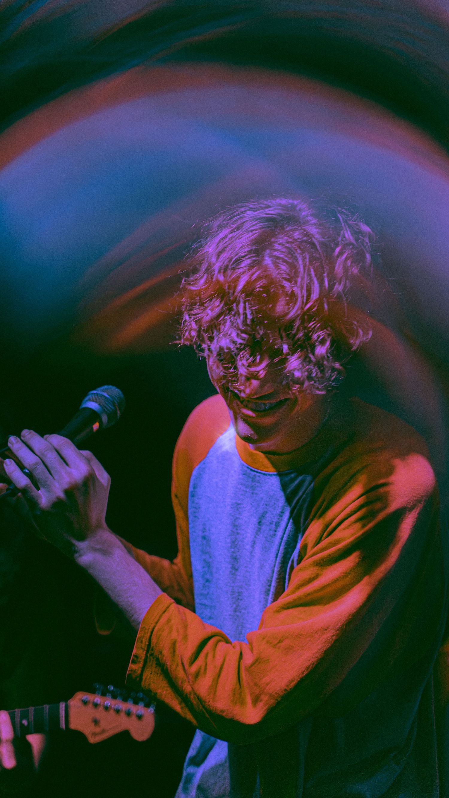 A musician performs on stage under vibrant purple and blue lighting while holding a microphone and guitar.