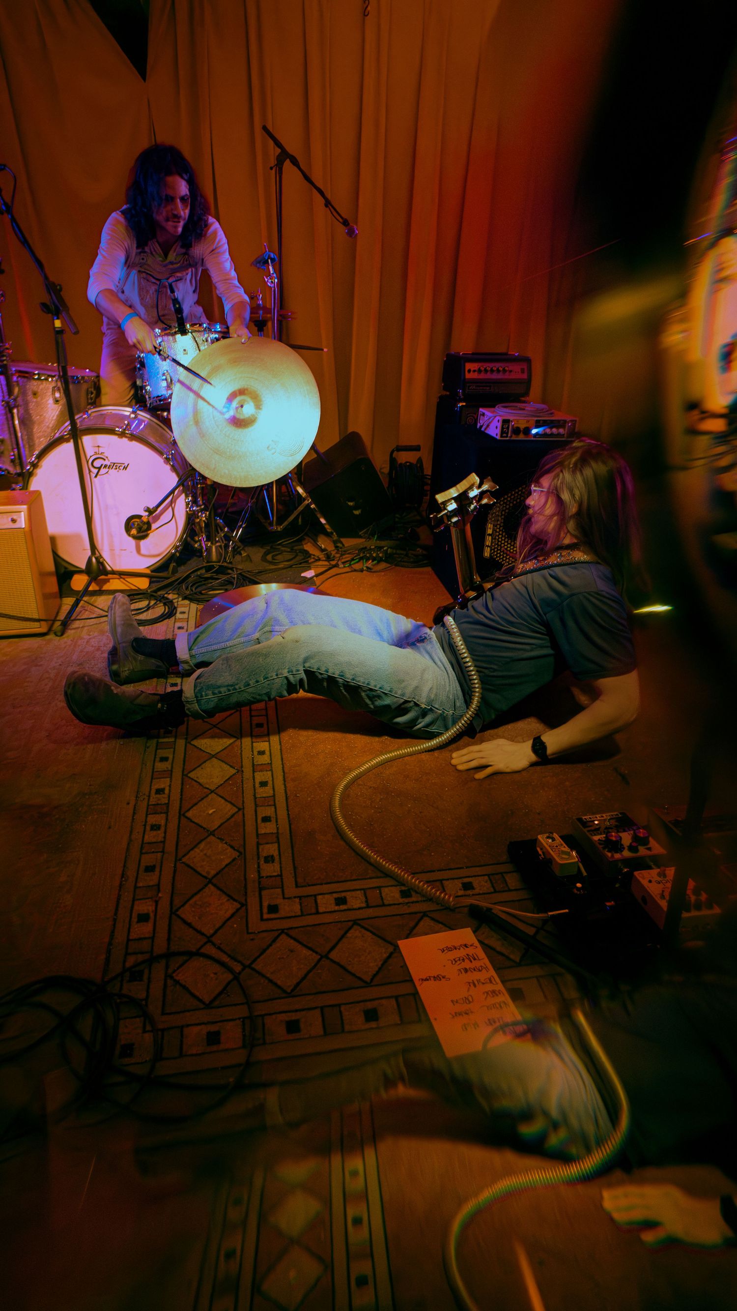 Musicians perform on the floor in a dimly lit room with an ornate rug creating a moody atmospheric vibe.